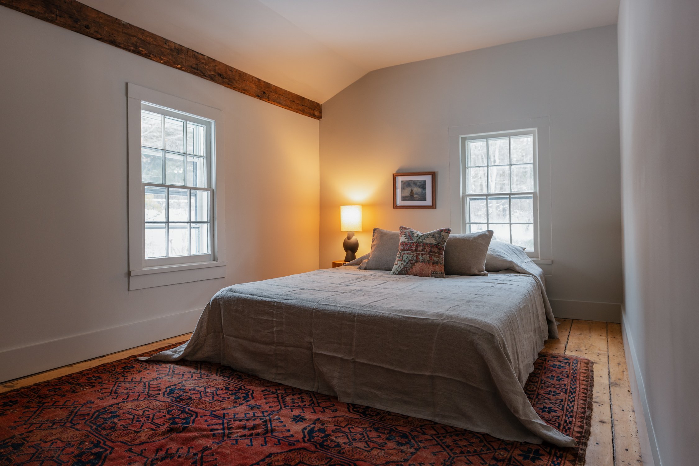 A bedroom with a bed covered in beige linens, three pillows, a small nightstand with a lamp, and two windows allowing natural light, with exposed wooden beam and a patterned rug on the wooden floor.