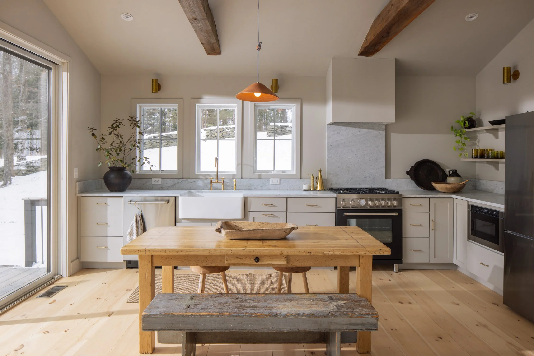 A modern kitchen with white cabinets, a white farmhouse sink, a black oven and stove, a black refrigerator, open shelves with dishes and decorative items, a large wooden dining table, and a sliding glass door leading outside to snow-covered ground.