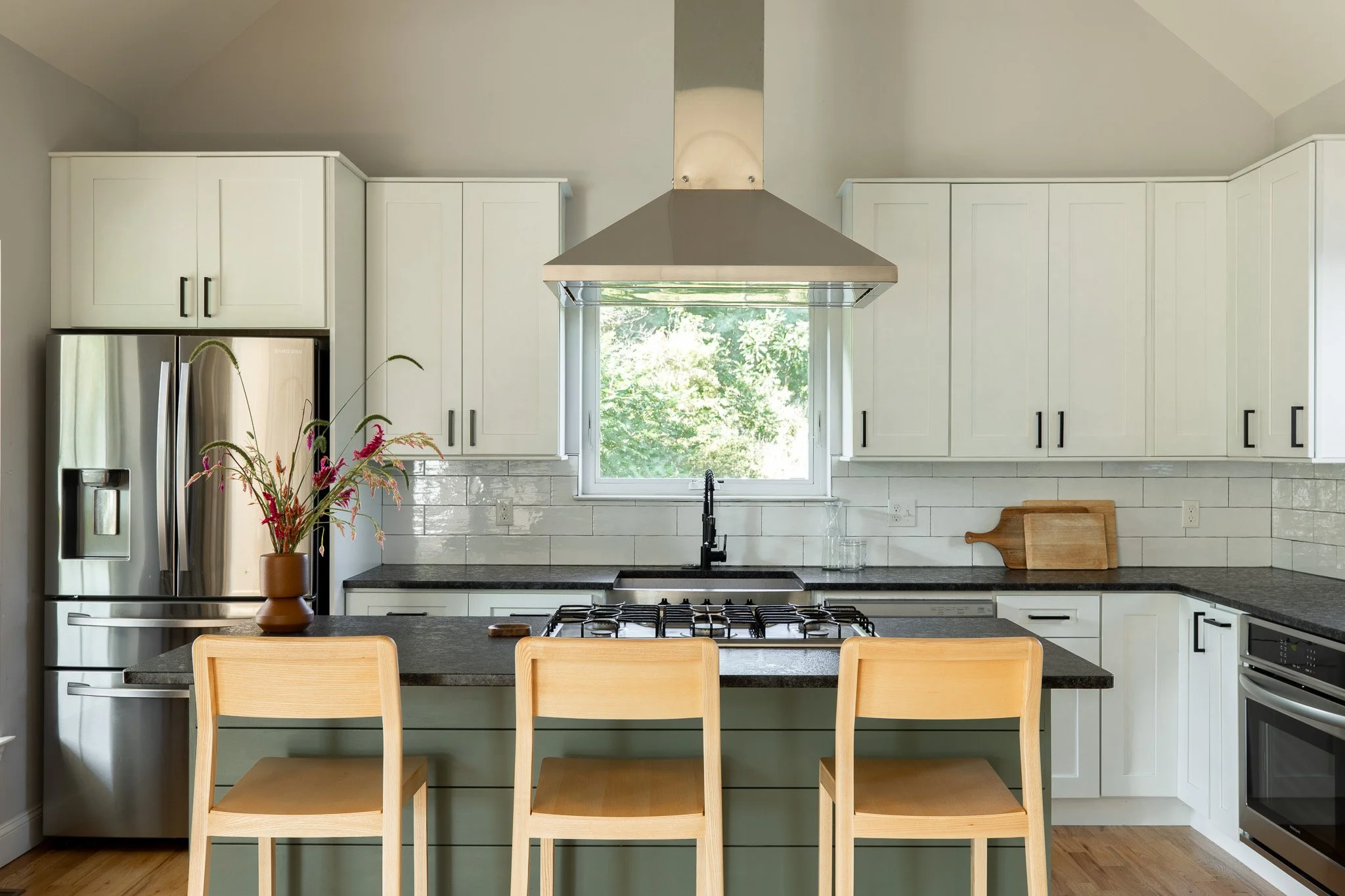 Modern kitchen with white cabinets, black countertop, stainless steel refrigerator, range hood, and a window overlooking greenery.