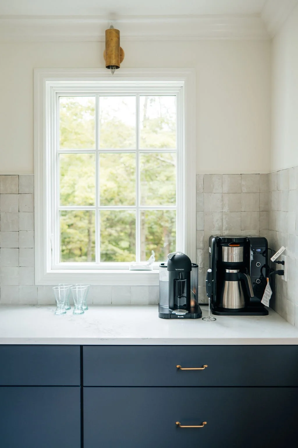 Kitchen countertop near a window with a coffee maker, a small espresso machine, and three empty glasses.