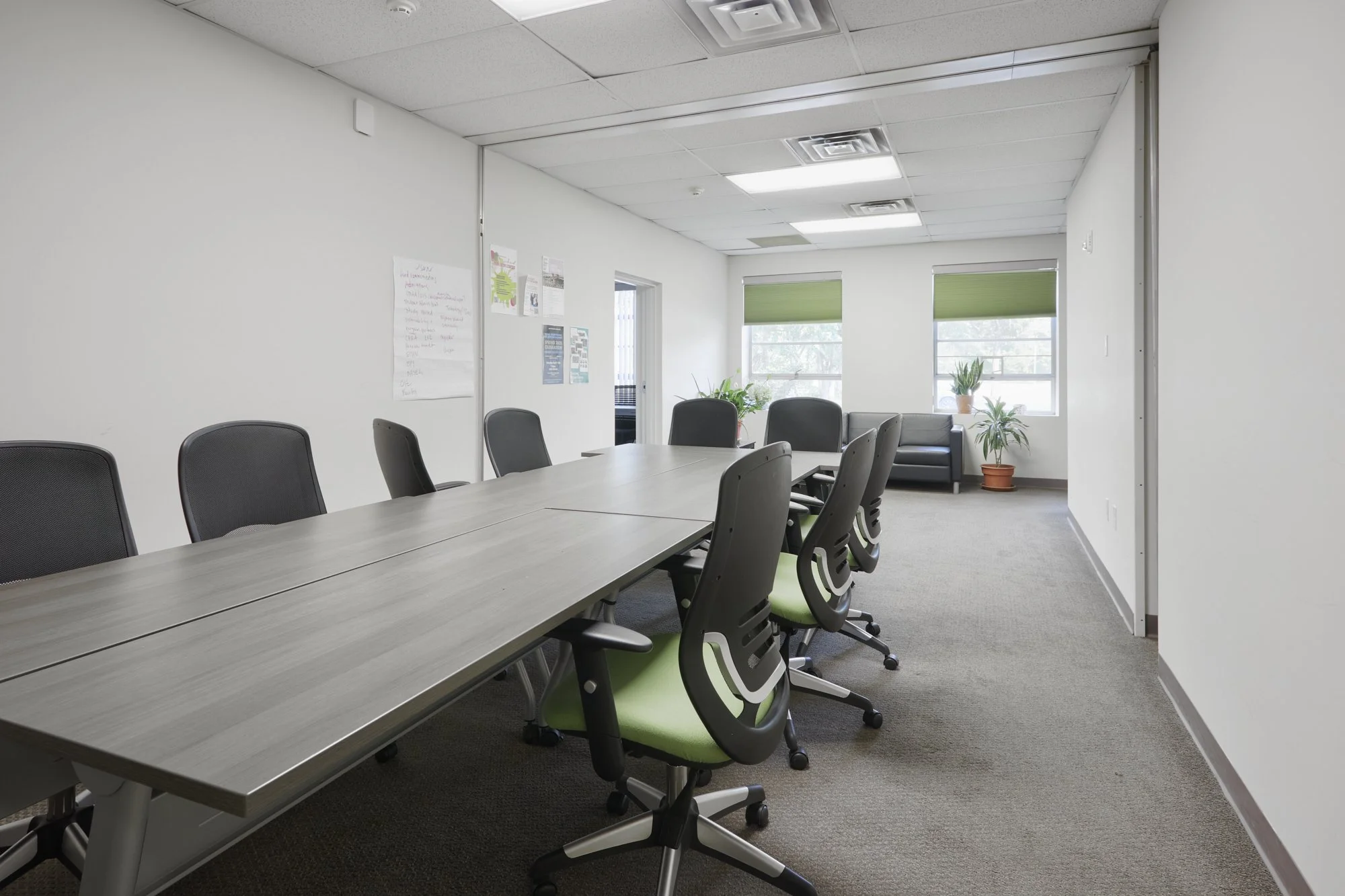 Empty conference room with a long table, black chairs with green seats, and large windows with green blinds, interior with beige carpet and white walls
