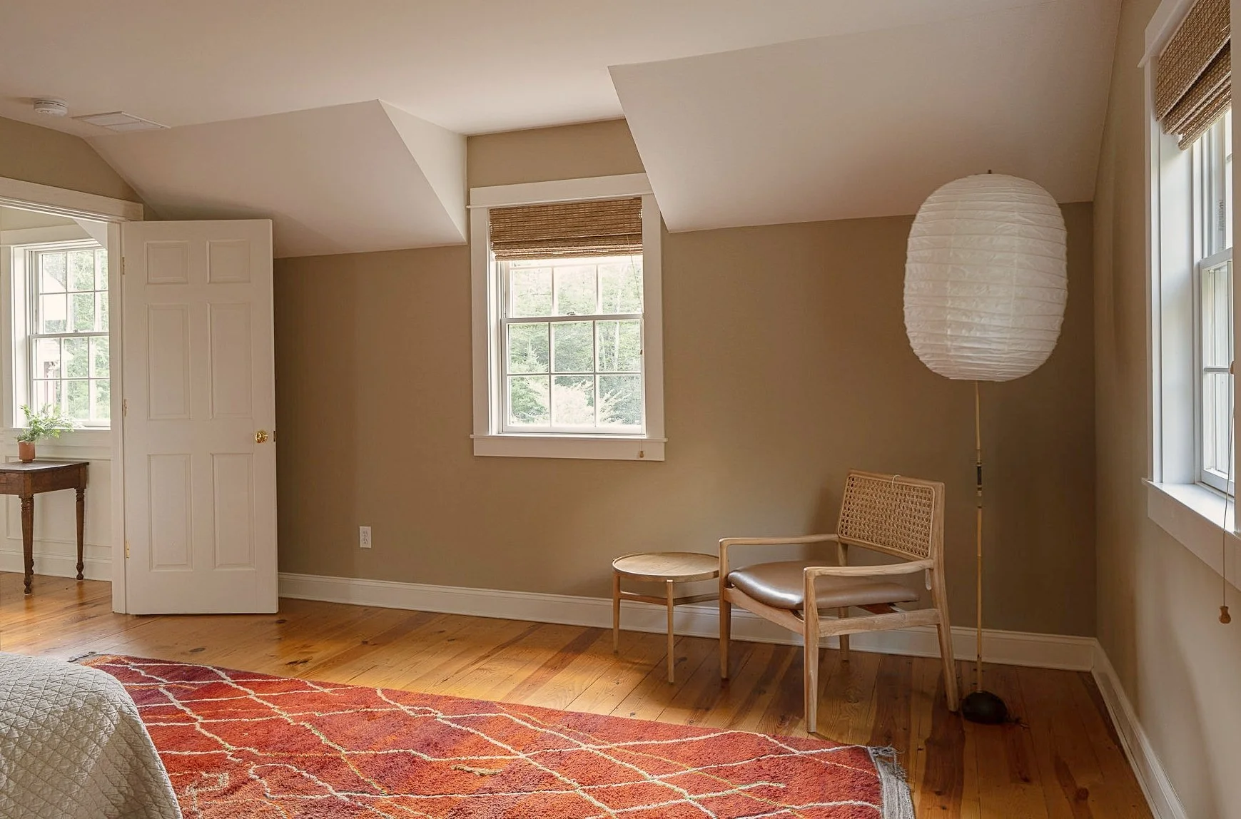 Empty bedroom with two windows, a beige wall, a wooden chair, a small side table, a paper lantern floor lamp, a red patterned rug, and a partially visible bed.
