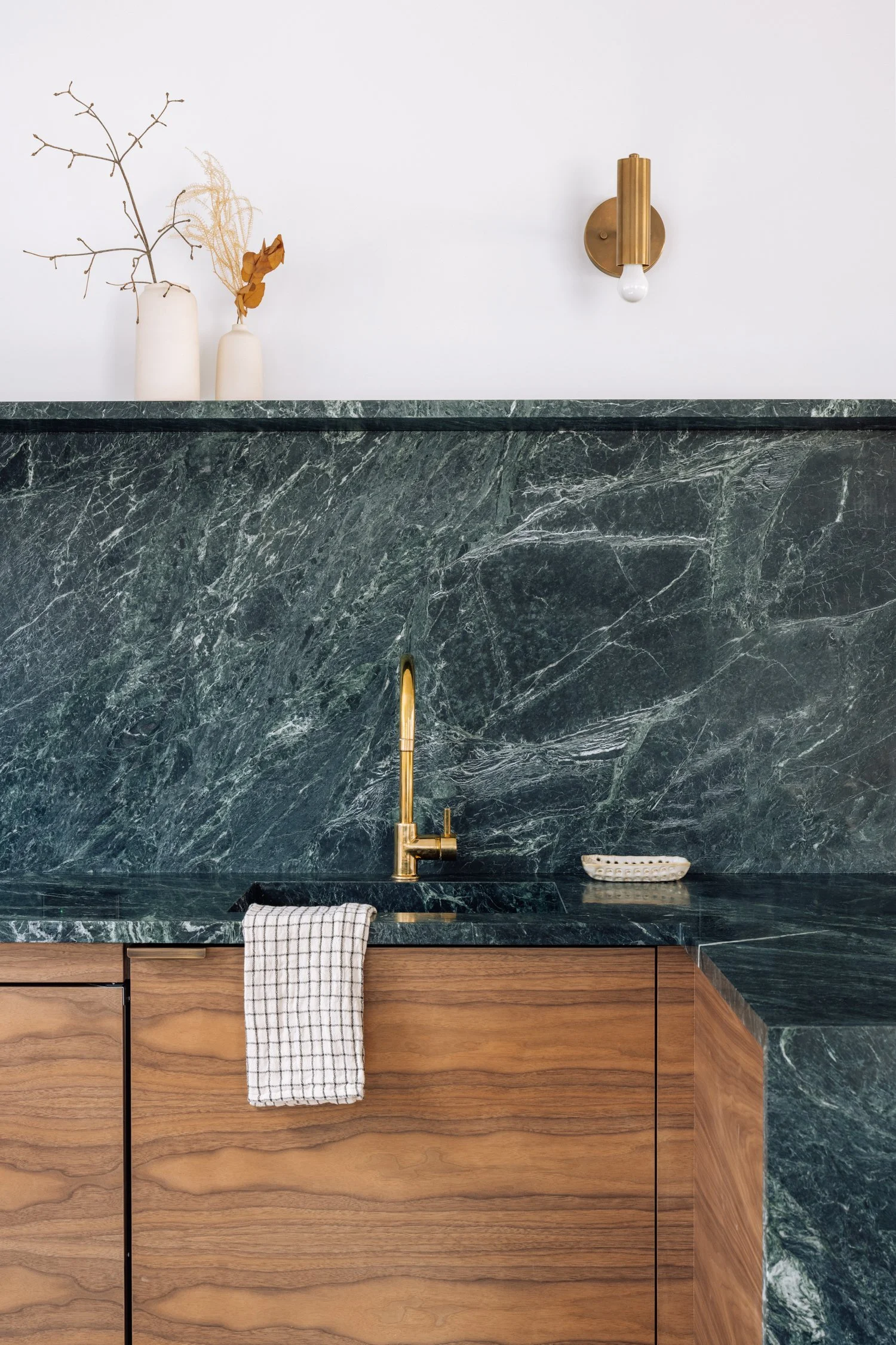 Modern kitchen with green marble countertops and backsplash, copper faucet, wooden cabinets, white wall with vases and wall light.