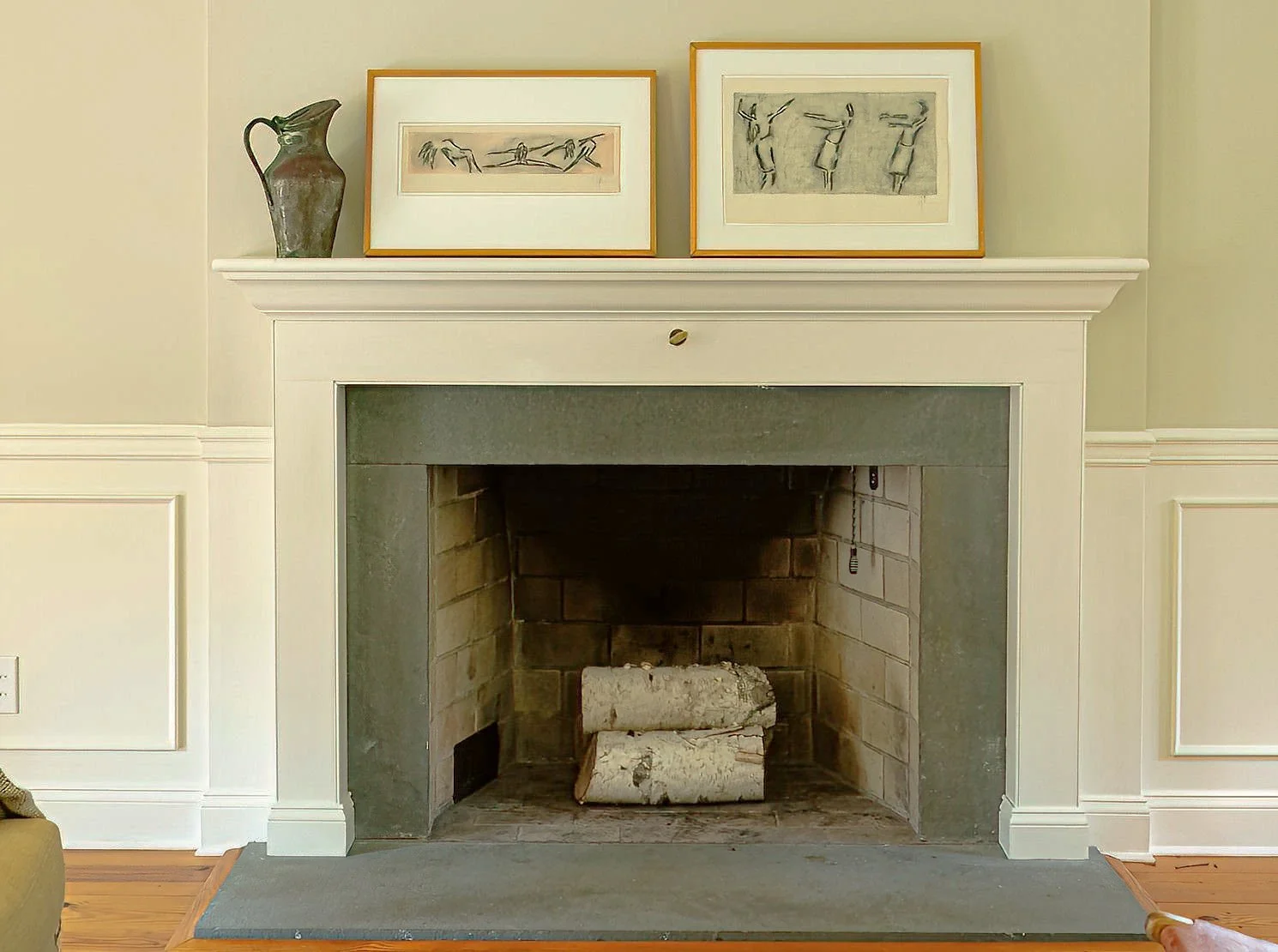 Interior view of a fireplace with two logs inside, above which sits a white mantel decorated with two framed artworks and a ceramic pitcher on the left.