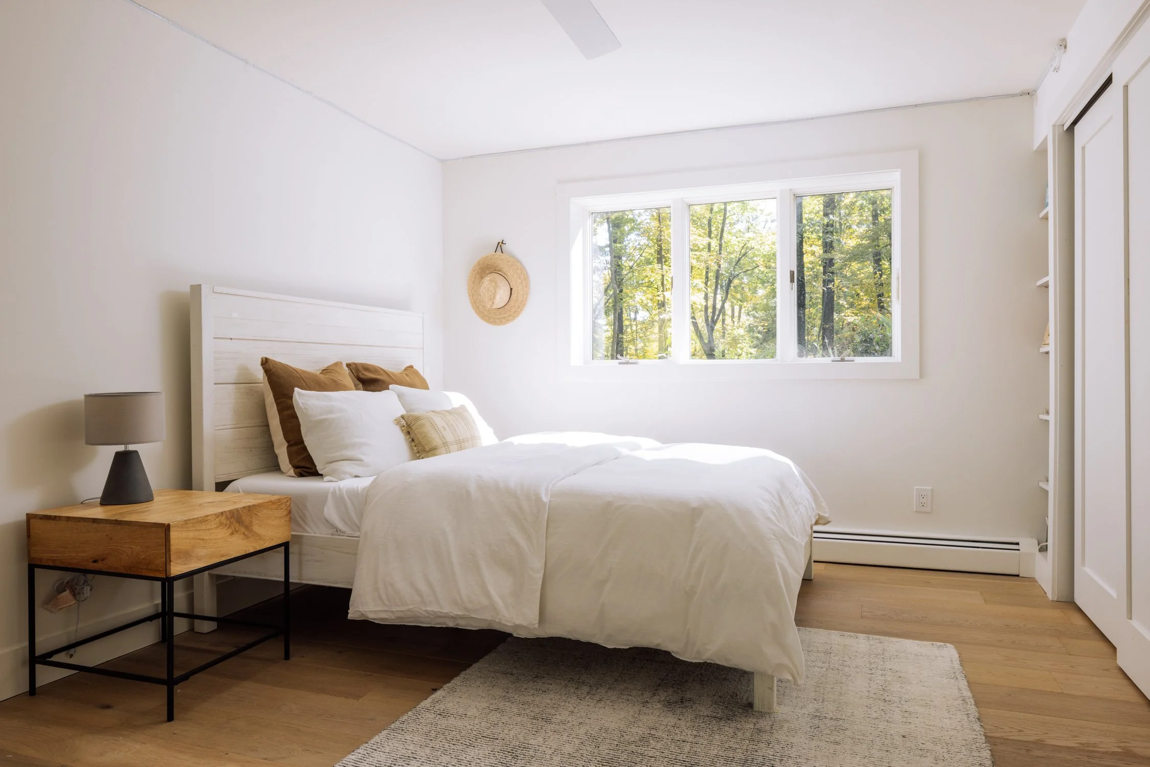 Bright bedroom with white walls, a bed with white and brown pillows, a wooden nightstand with a lamp, a window showing trees outside, and a beige rug on the wooden floor.