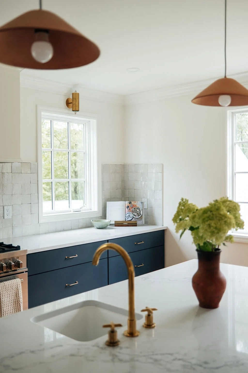 Modern kitchen with white marble island, brass faucet, blue cabinets, two pendant lights, window, beige backsplash, and decorative items including a green bowl, a book, and a vase with green flowers.
