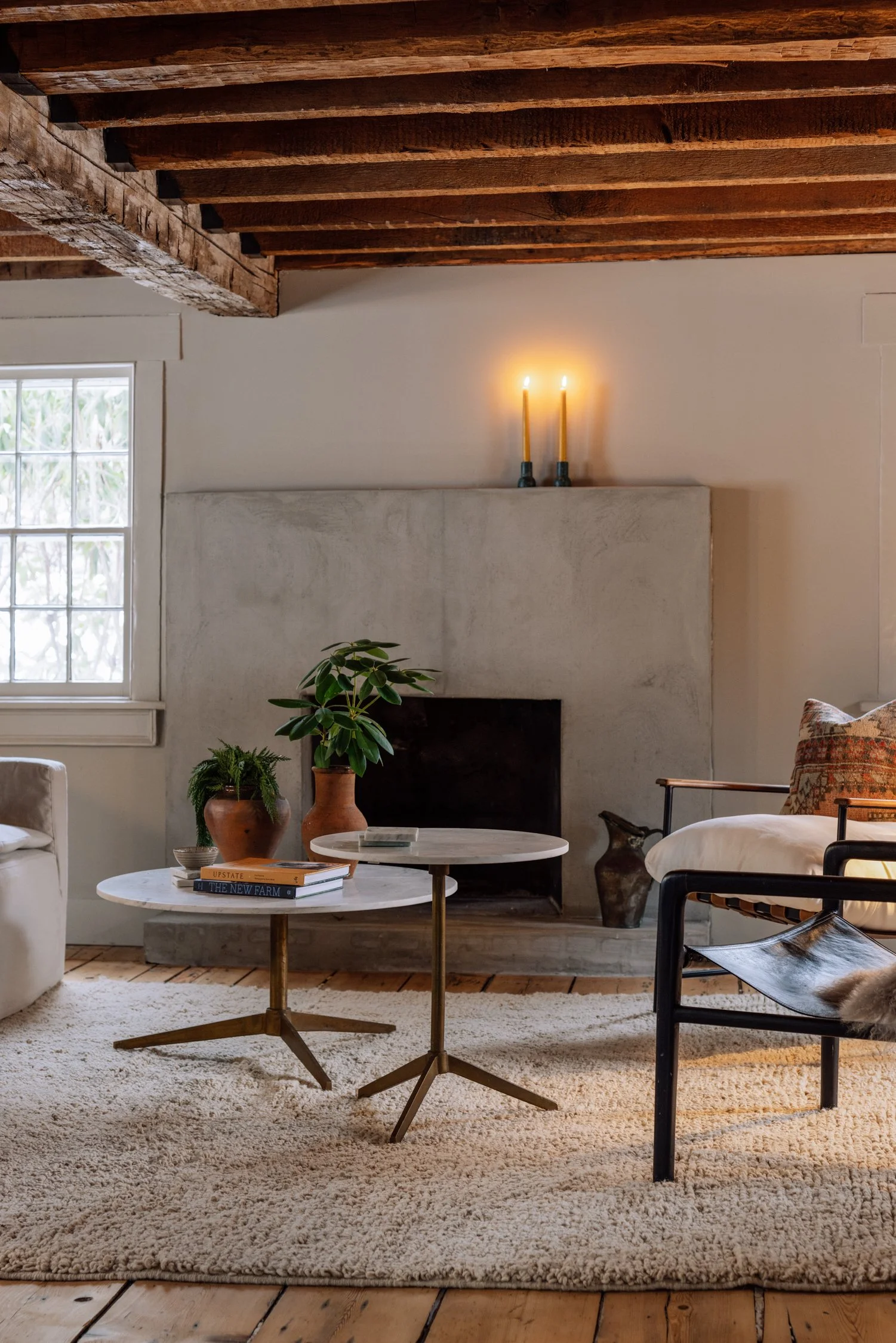 Living room with white walls and wooden ceiling beams, a fireplace, a window with grid panes, two marble-topped side tables with brass legs, a beige rug, a white sofa, a black armchair with a cushion, potted plants, and lit candles on the mantel.