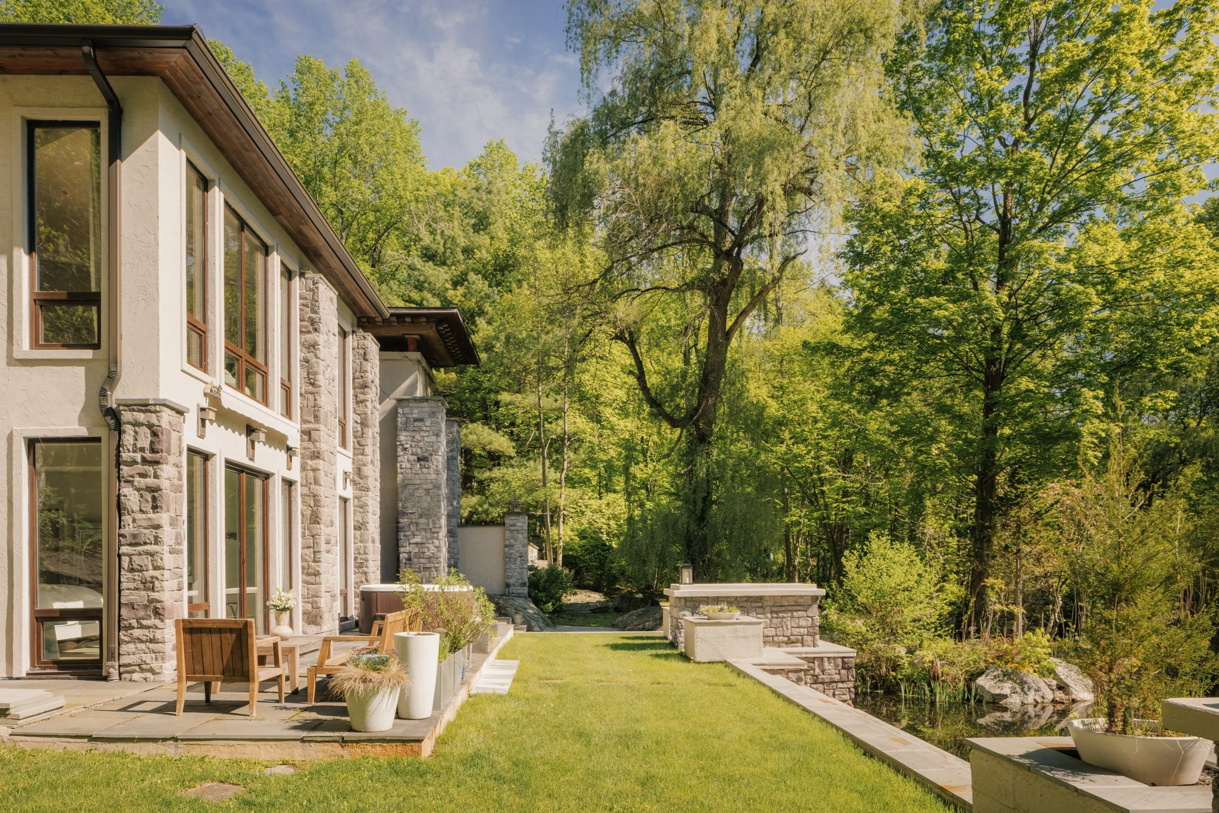 Modern house with a stone and stucco exterior, surrounded by lush green trees, with a patio and garden area.