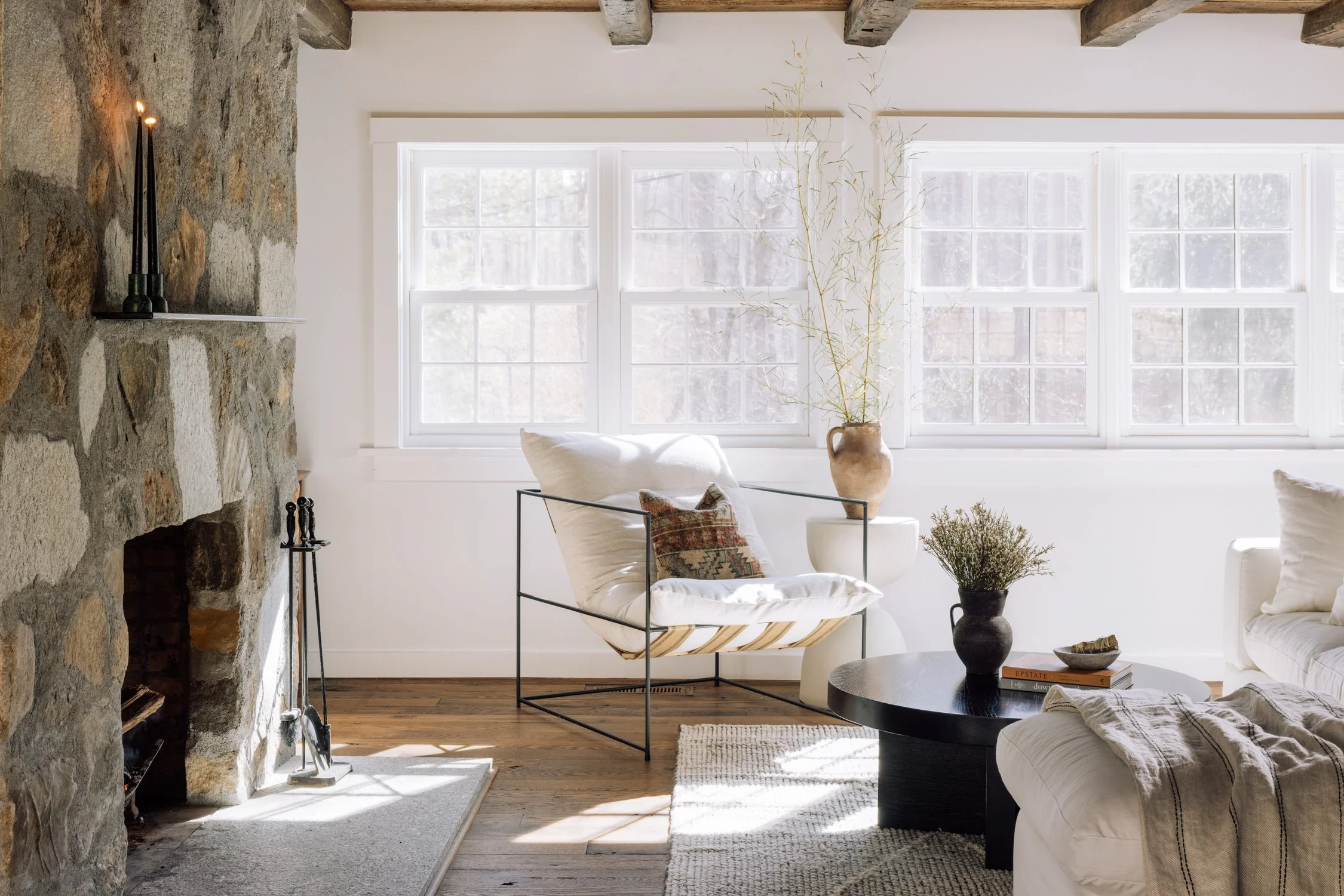 Living room with white walls, wooden ceiling beams, a stone fireplace, large windows, and furniture including a white chair with a pendant-style pillow, a black round coffee table with books and vases, and a beige sofa. Decor includes a tall vase wit