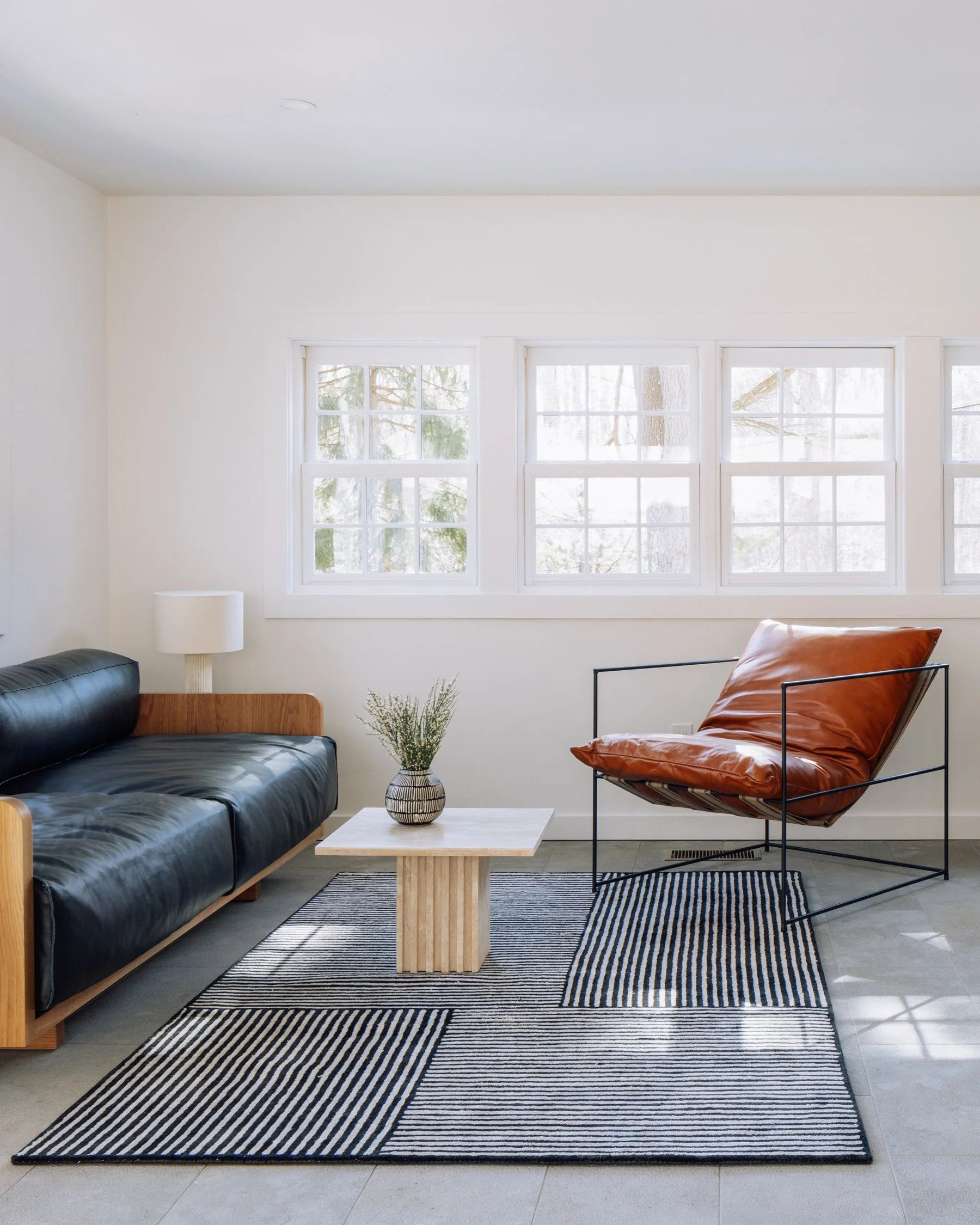A minimalist living room featuring a black leather sofa with wooden accents, a white table with a small vase of flowers, a modern brown leather lounge chair with a black metal frame, and a striped black and white rug, with four large windows letting 