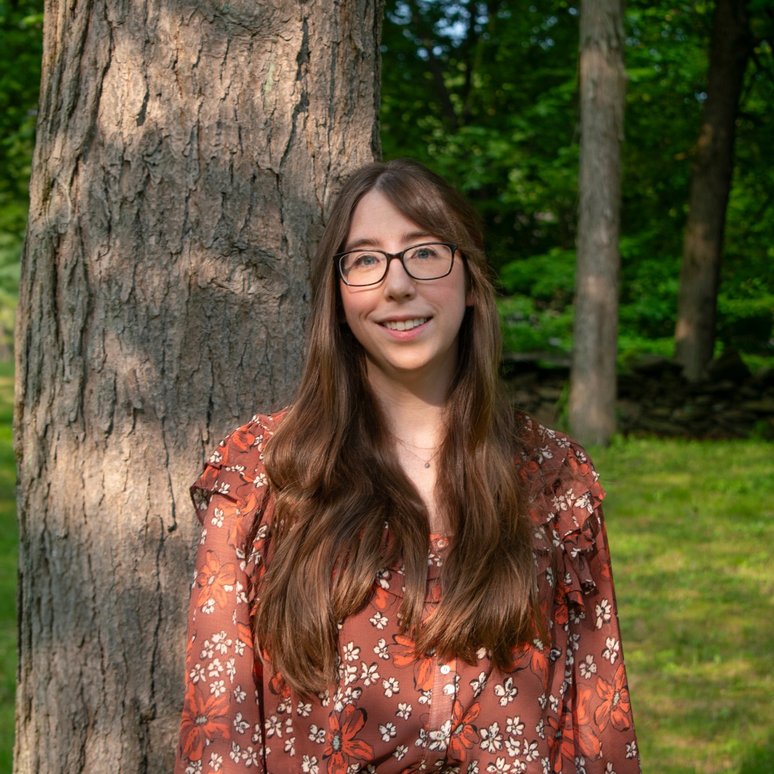 A young woman with long brown hair, glasses, and a floral blouse standing outdoors next to a large tree with a lush green forest background.