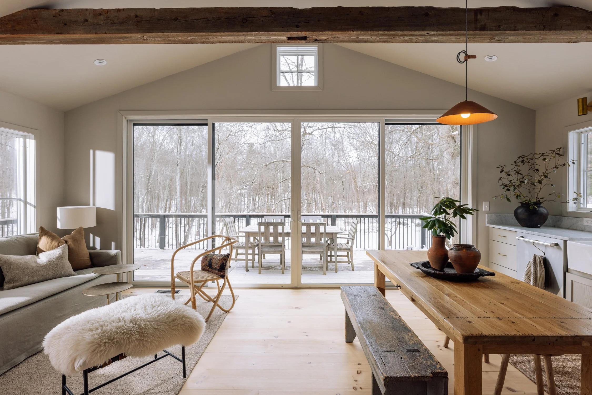 Living room with large sliding glass doors leading to a snow-covered deck and a wooded outdoor area, featuring a beige sofa with pillows, a sheepskin footstool, a wooden dining table with a bench, and potted plants.