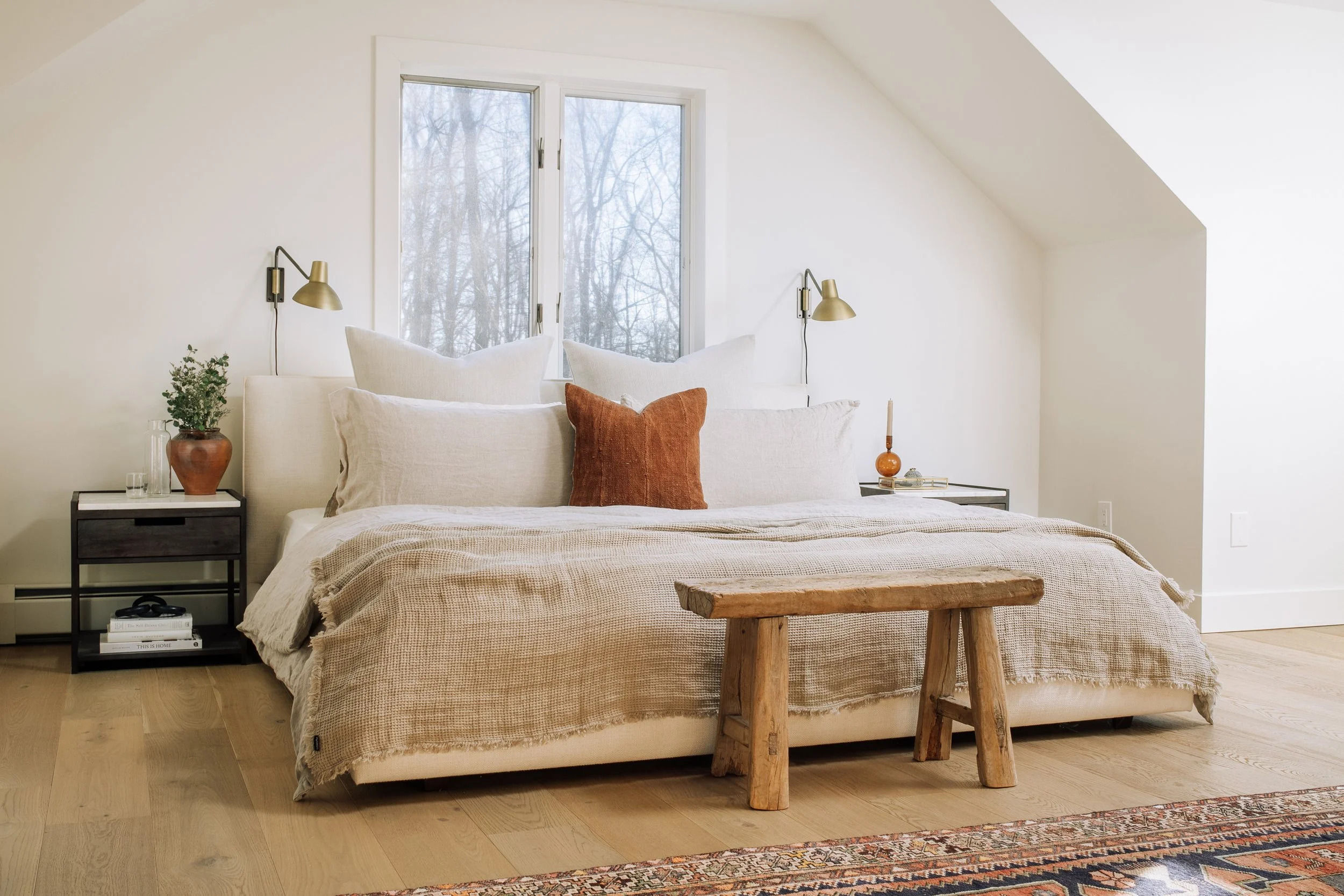 A cozy bedroom with a white bed, multiple white pillows, and a rust-colored accent pillow. The bed is beneath a window with a view of leafless trees outside. Two black side tables with decorative items and wall-mounted gold lamps are on either side o