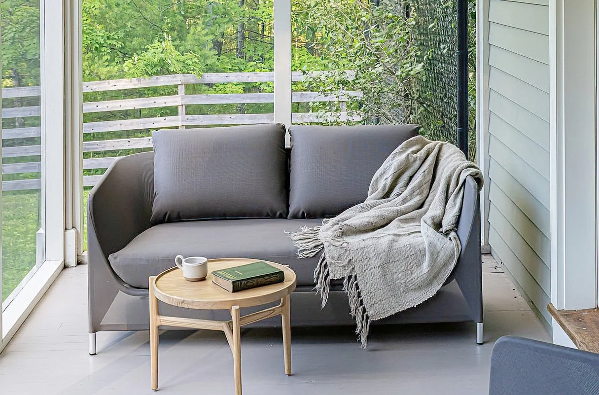 A cozy porch area with a gray sofa, a light wood side table with a cup and left book, and a beige blanket draped over the right arm of the sofa, overlooking a lush green backyard with trees and a wooden fence.