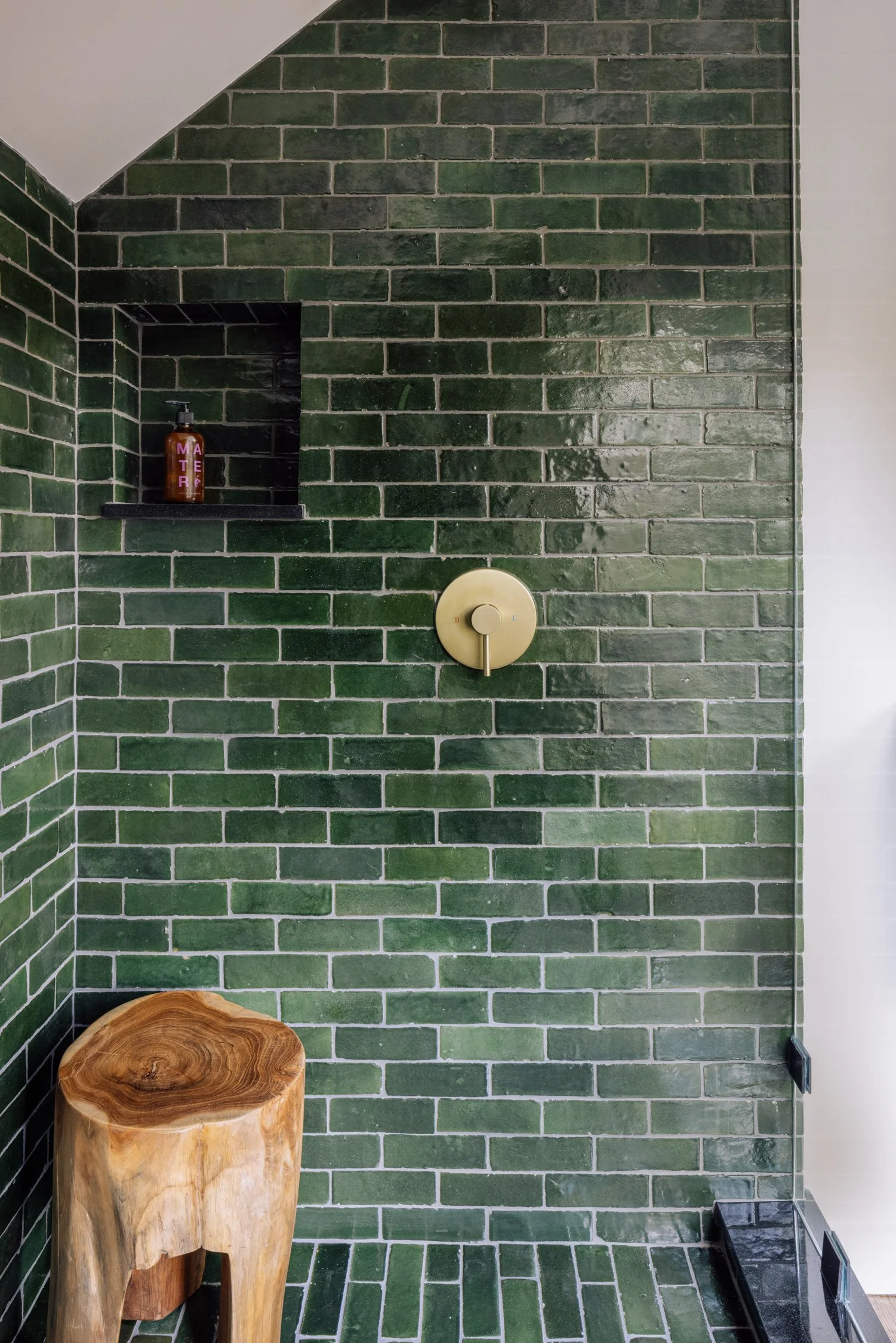 Green tiled shower with a built-in shelf holding a brown bottle labeled "MATTER," and a brass shower control handle, next to a natural wood stool.