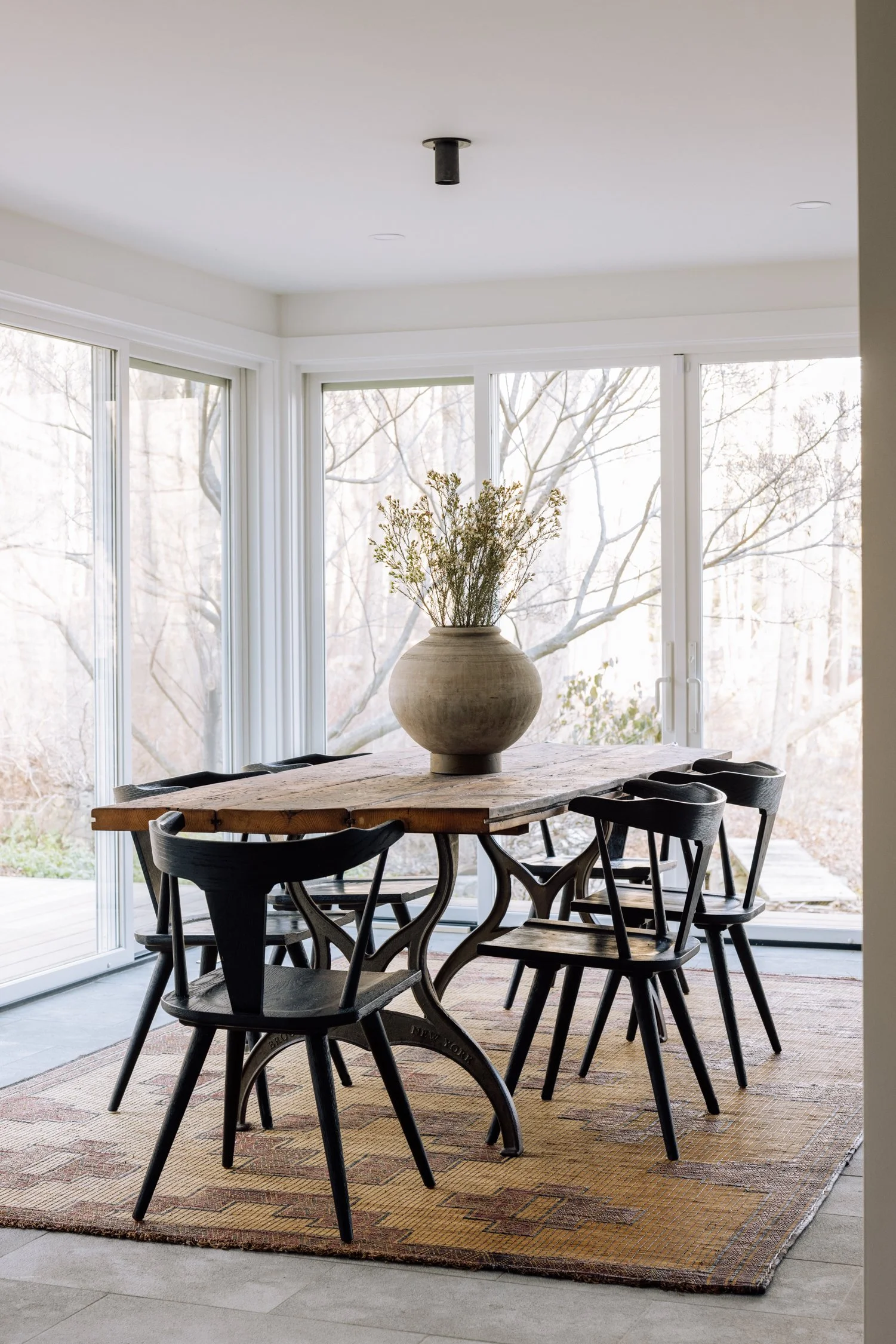 Dining room with large windows, a wooden table, six black chairs, a large vase with dried flowers, and a patterned rug.