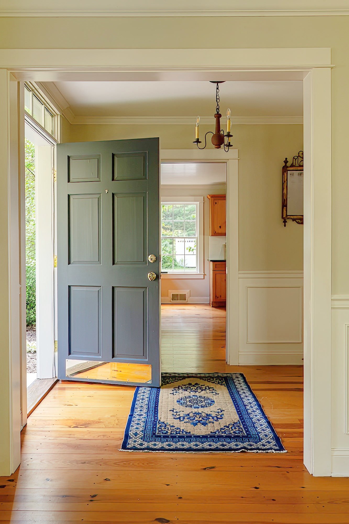 Interior view of a house with a front door open, revealing a hallway with a wooden floor, a blue rug, and a chandelier hanging from the ceiling.