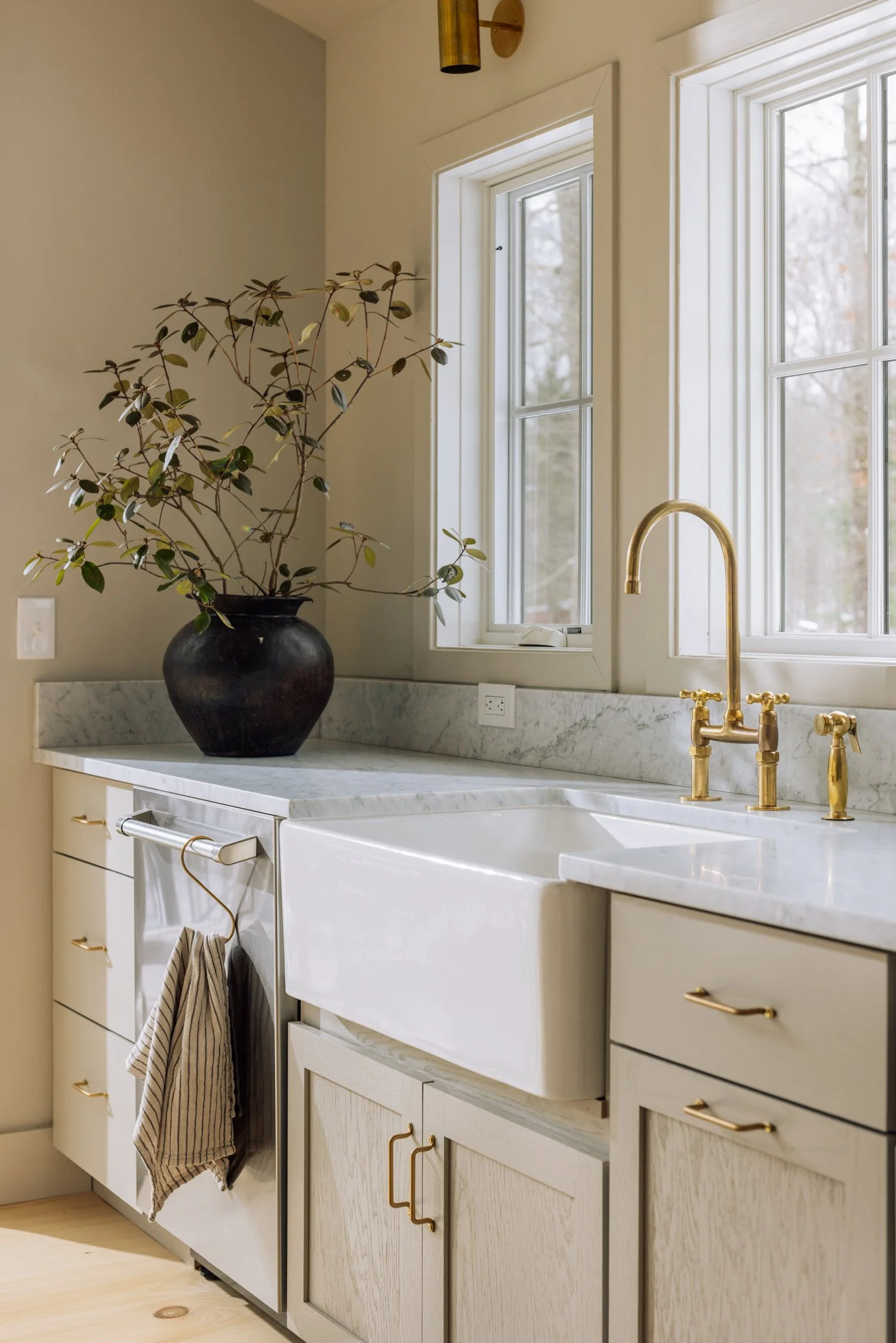 Kitchen with white marble countertop, farmhouse sink, brass faucet, large window, and a potted plant with dark oval leaves on the counter.