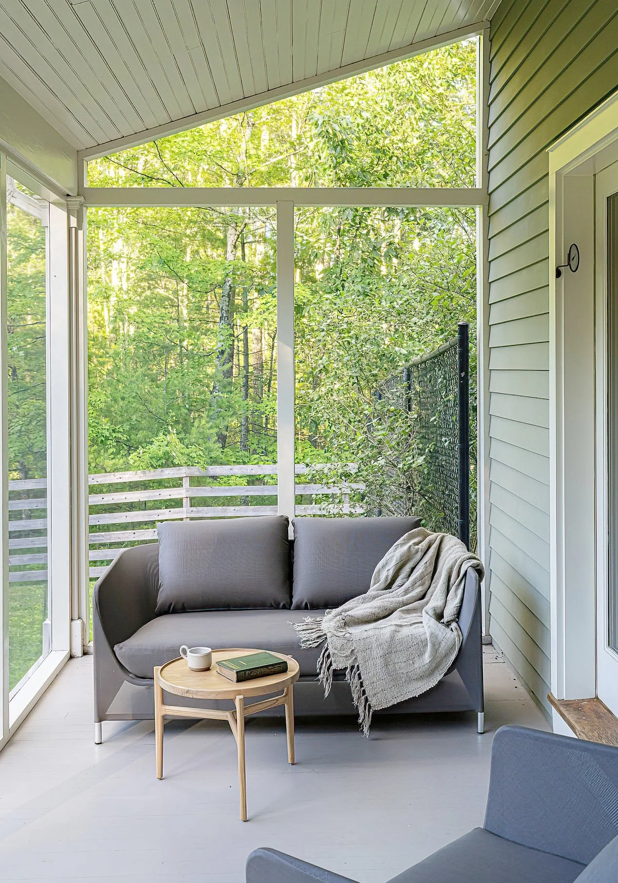 Screened porch with gray sofa, beige throw blanket, small wooden side table with a book and mug, overlooking green trees and a wooden fence.