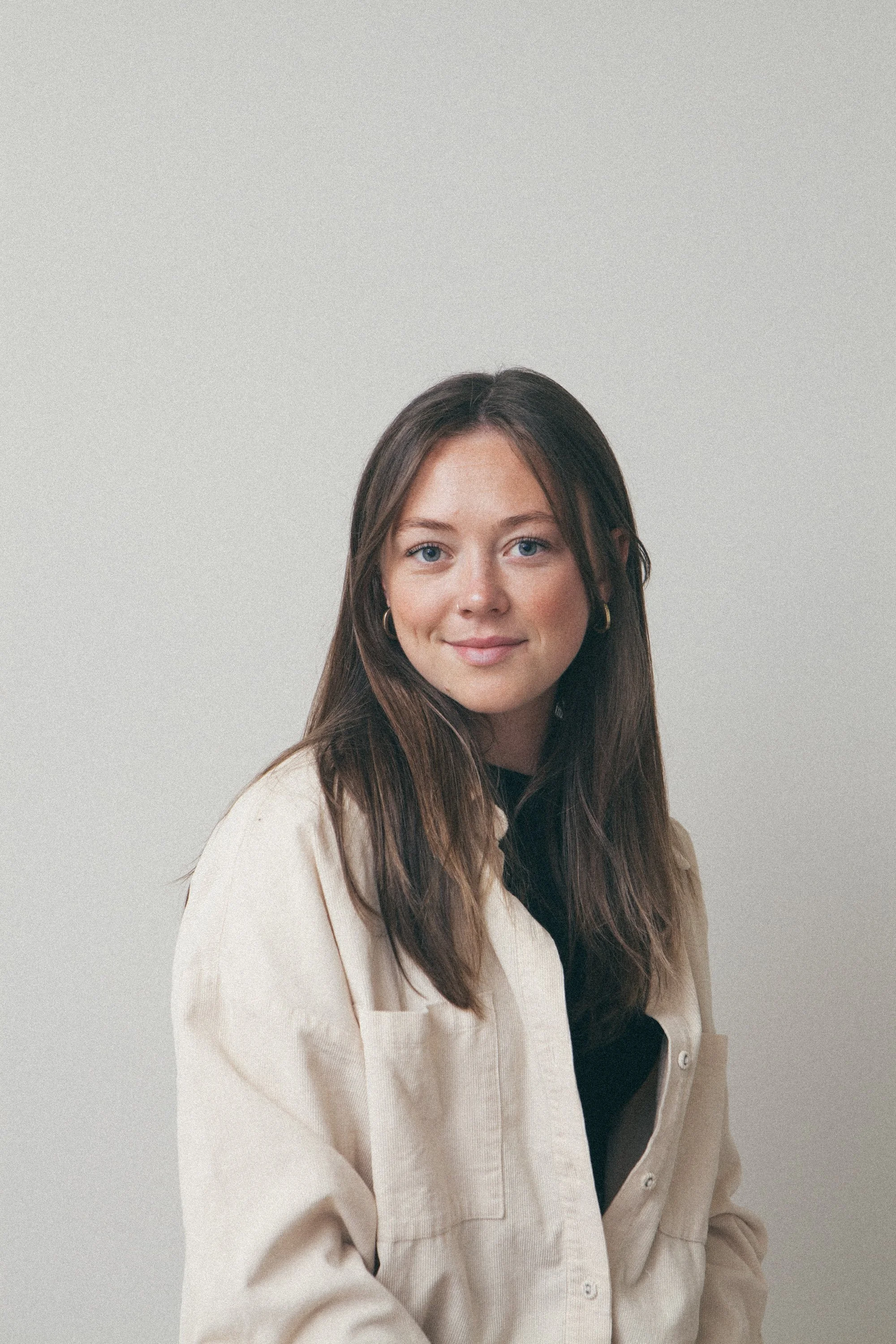 A woman with long brown hair, blue eyes, and a nose piercing, smiling and standing in a forest, wearing a light blue blazer over a white shirt.