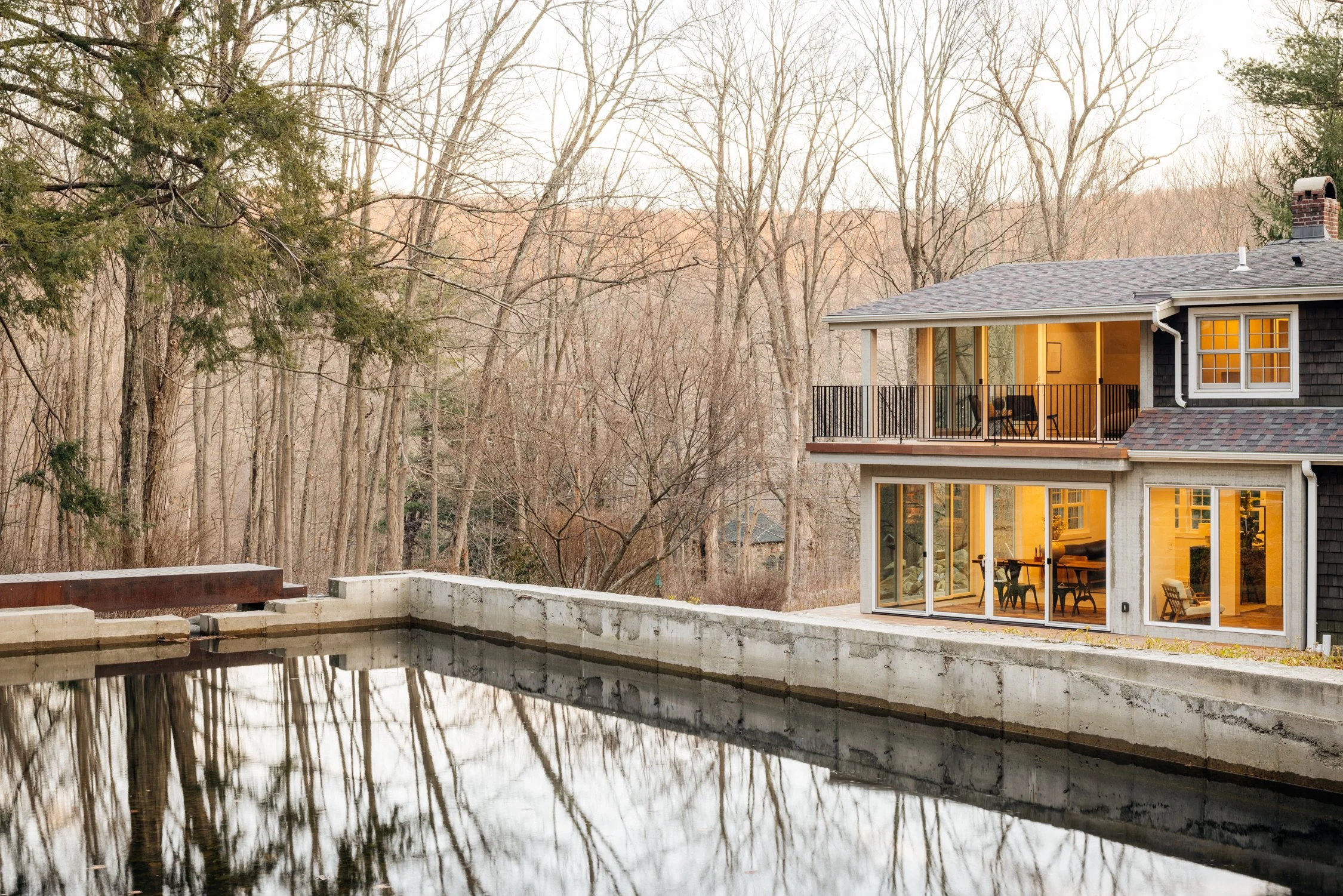 House with a screened porch and large glass doors, overlooking a pond with trees in the background at dusk.