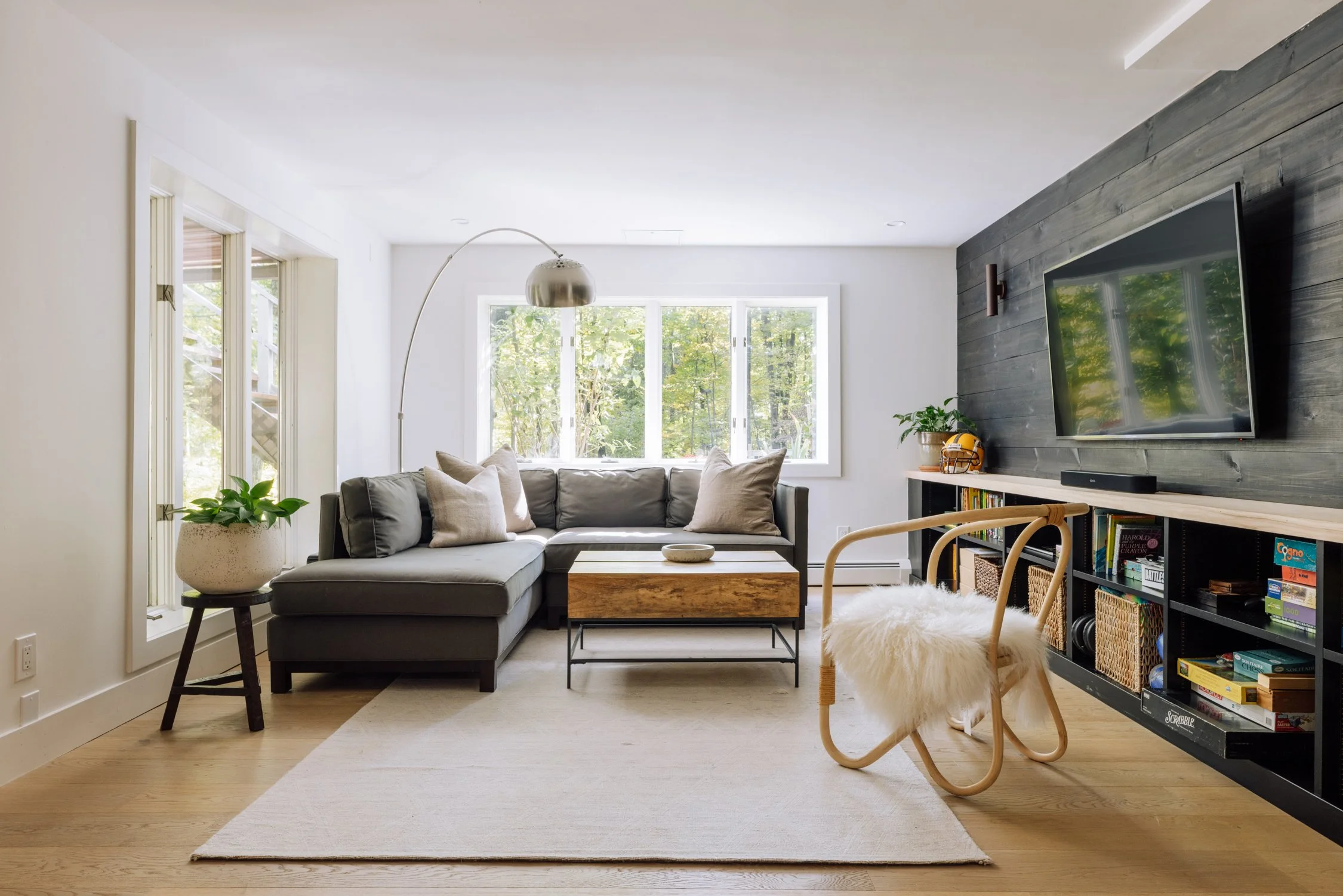 Modern living room with gray sectional sofa, wooden coffee table, white walls, wooden floor, large window with trees outside, black bookshelf with baskets and books, wall-mounted tv, white and wooden accents, and a rocking chair with a white fur cush