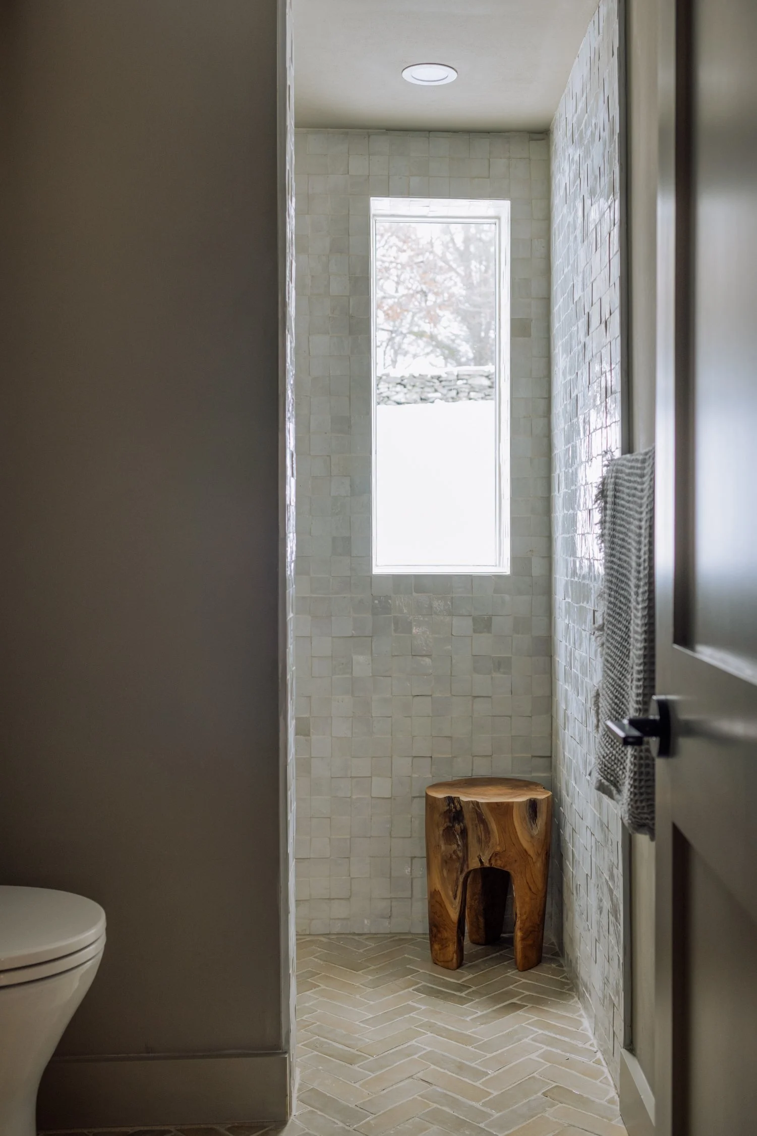 A minimalist bathroom with a tall narrow window, tiled walls, and a wooden stool on a herringbone tile floor.