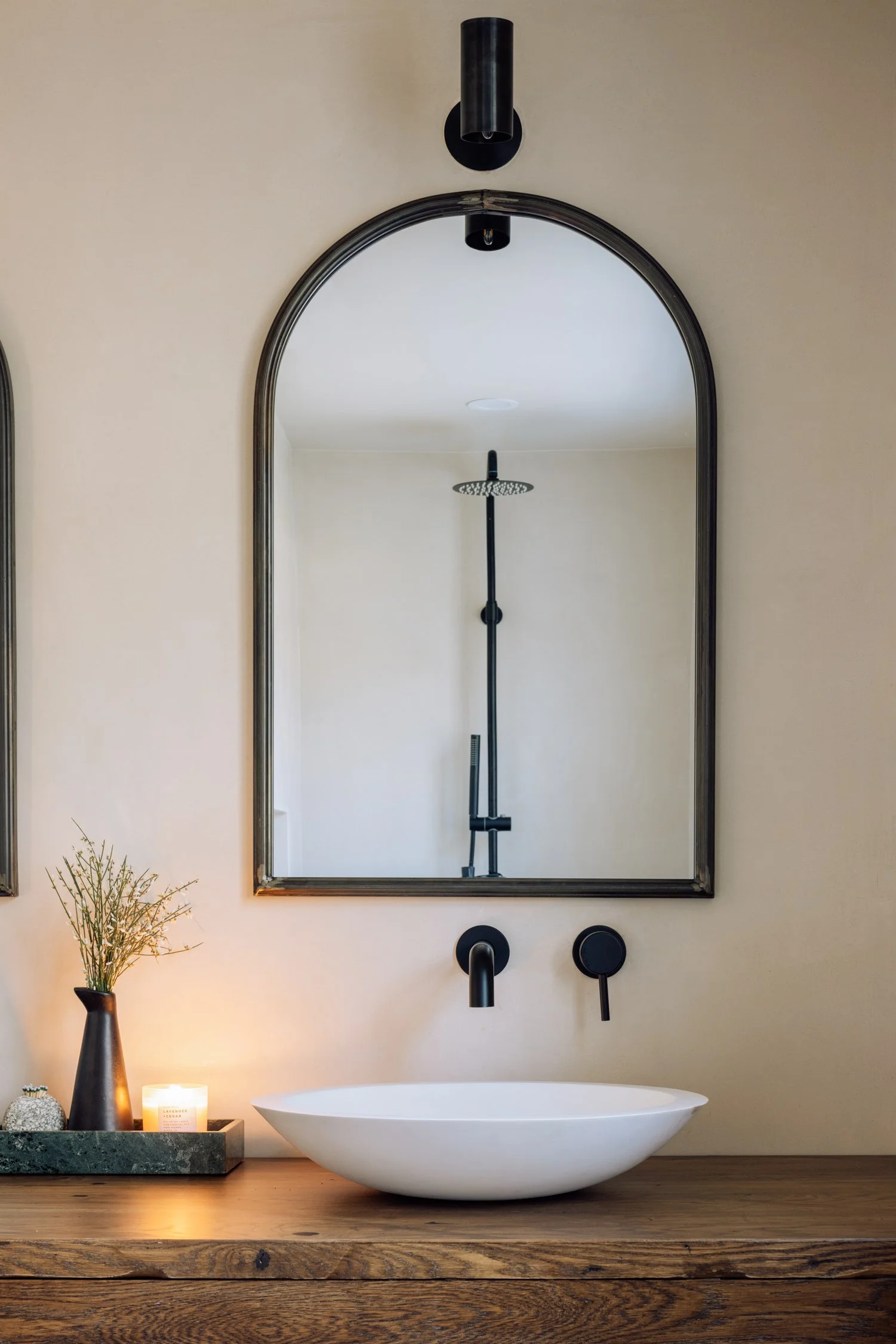 Bathroom with a white vessel sink on a wooden countertop, a large arched mirror, wall-mounted faucet, a small black vase with dried flowers, a lit candle, and a modern black showerhead in the reflection of the mirror.