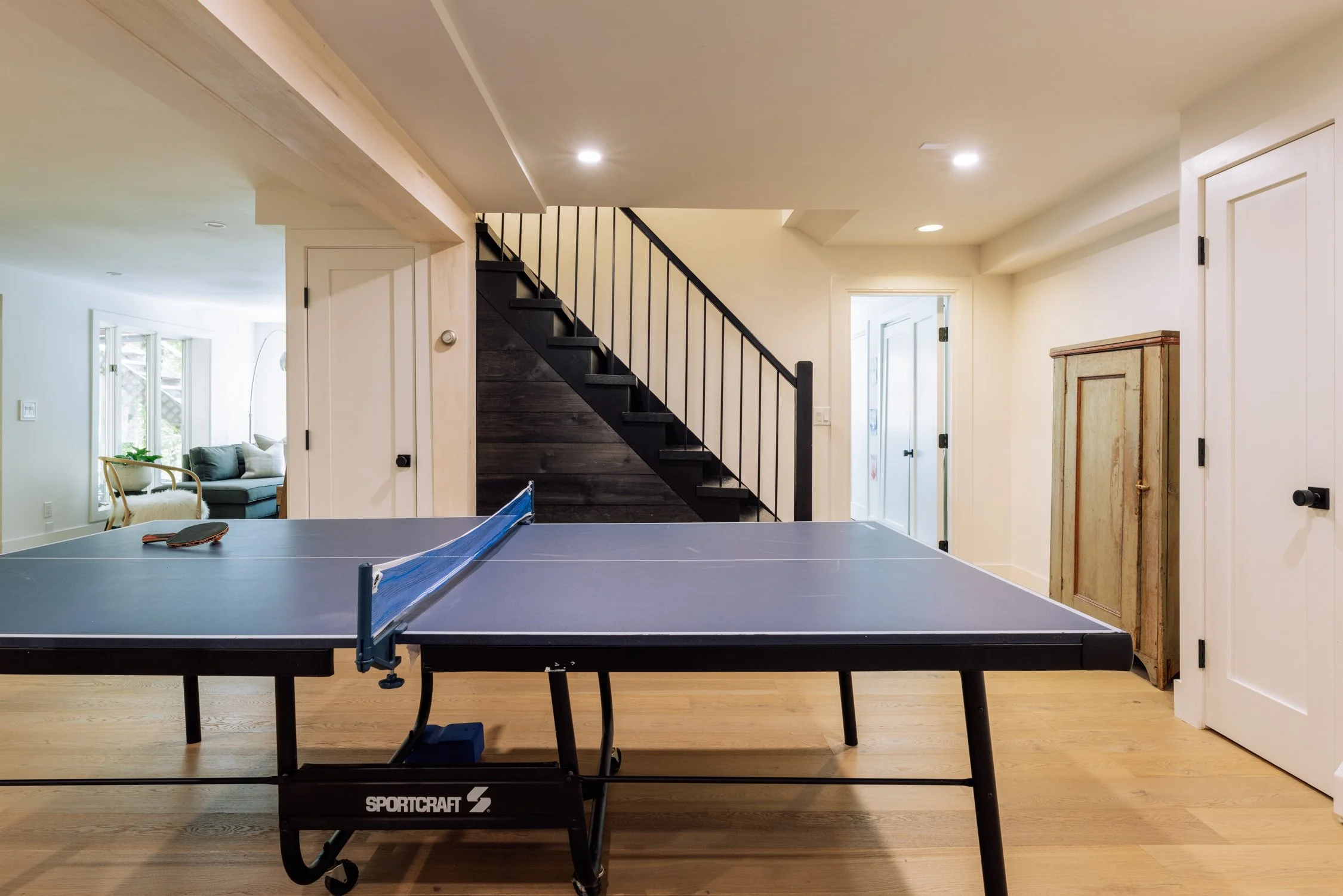 Indoor room with ping pong table, staircase, and wooden cabinets, with a living area visible through an open doorway.