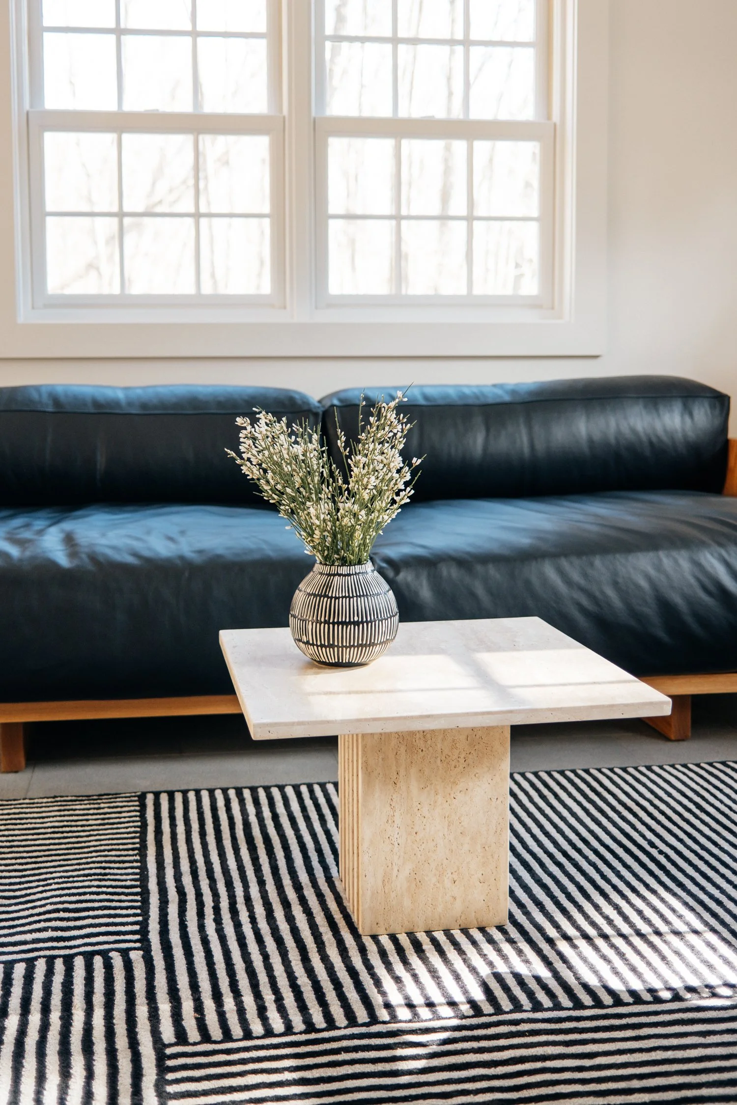 Interior of a living room with a black leather couch, a small cream-colored marble-topped table with a patterned vase of white flowers, a black and white striped rug, and large windows with white frames letting in natural light.