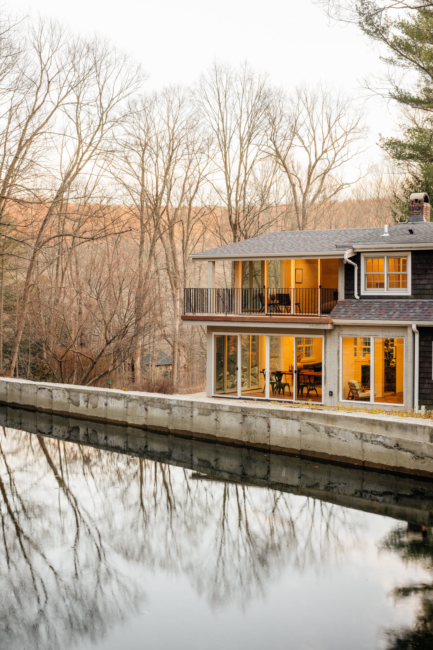 A house with large glass sliding doors and windows, overlooking a waterway, with leafless trees in the background, during evening or dusk.