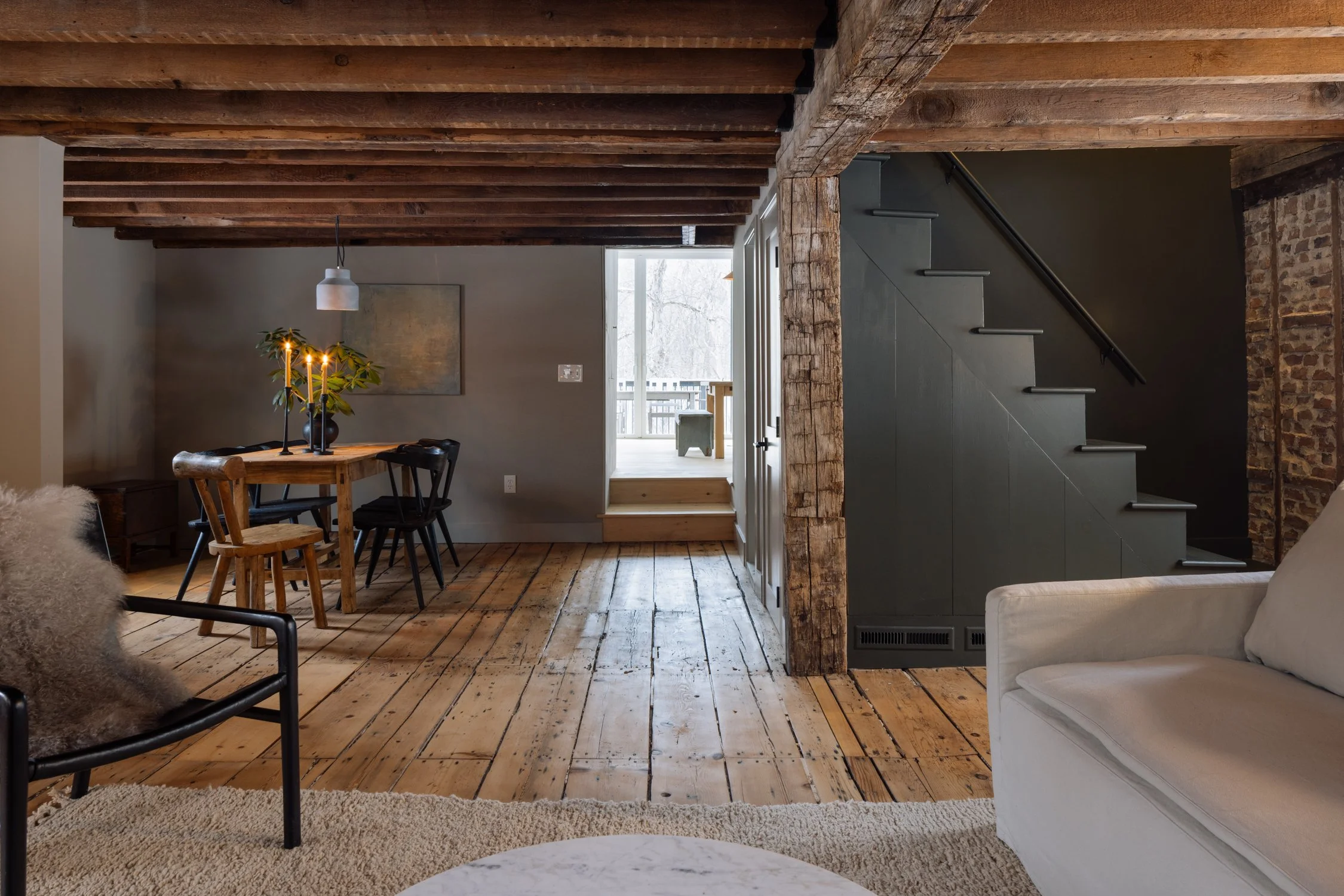 Interior of a rustic living room featuring exposed wooden beams, a wooden dining table with chairs, a stairway, and part of a sitting area with a sofa and an armchair on a beige rug.