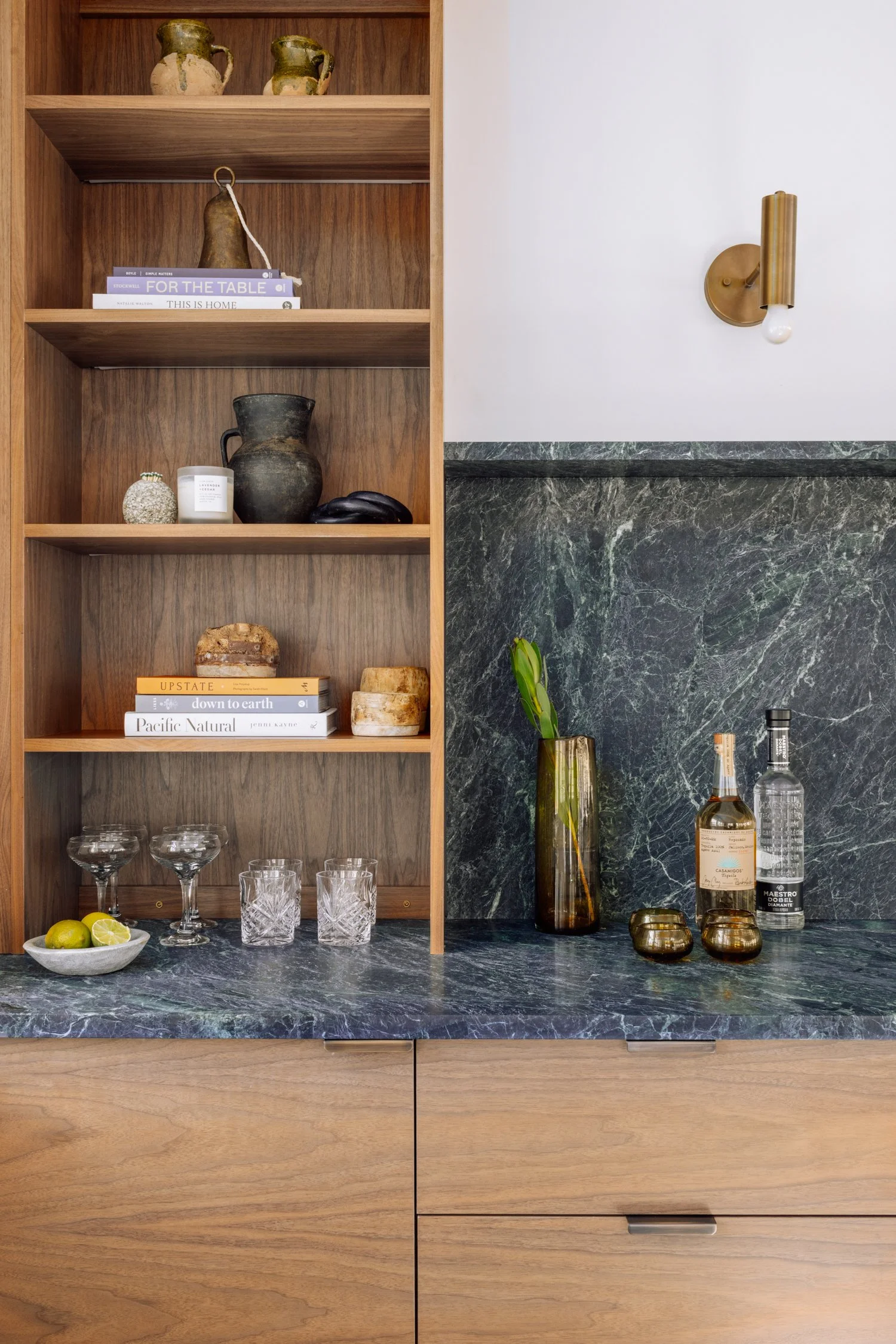 A wooden kitchen shelf with decorative vases, books, and glassware, next to a marble countertop with a tall glass vase containing greenery, bottles, and small decorative items.