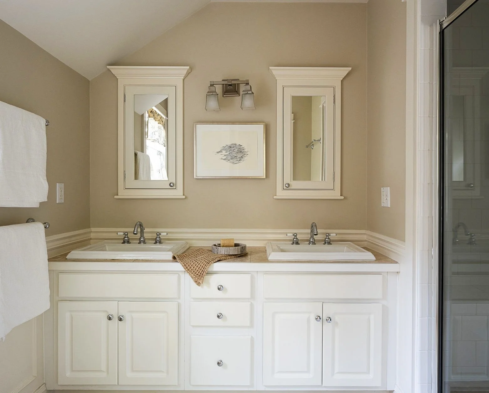 Bathroom vanity with two white sinks, mirror cabinets, a framed picture, and a towel rack on the left, with a shower on the right.