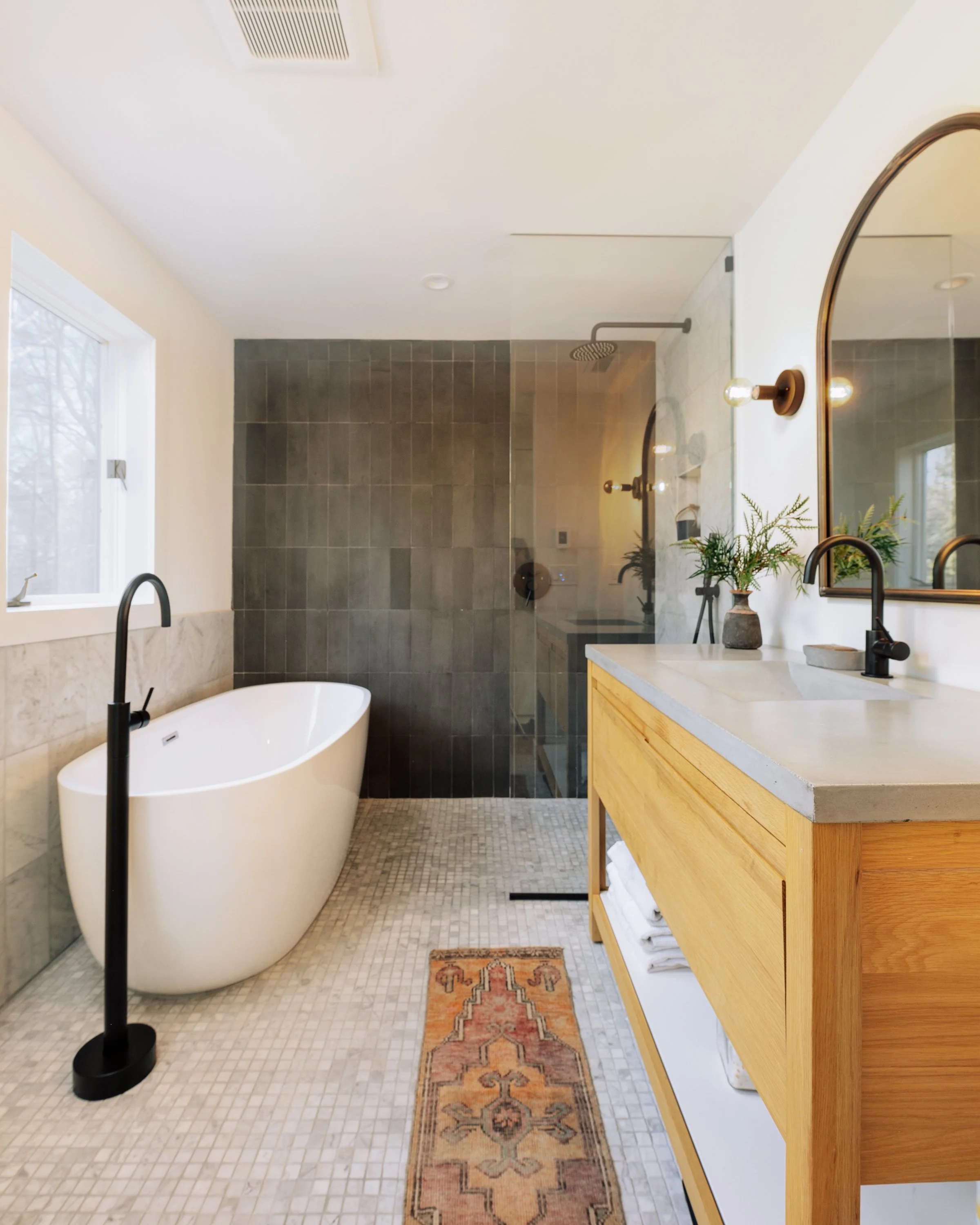 Modern bathroom with a white freestanding bathtub, black floor-mounted faucet, glass-enclosed walk-in shower with dark gray tiles, light wood vanity with concrete countertop, large mirror, potted plant, and small colorful rug on tiled floor.