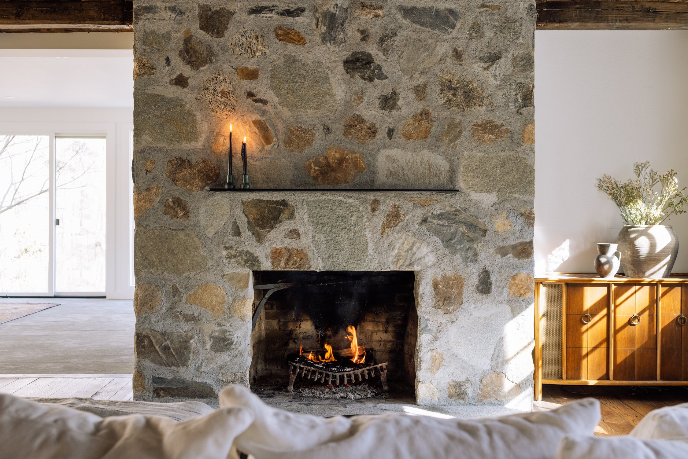 Stone fireplace with a lit fire, a black shelf with two lit candles above, a wooden sideboard with vases and a large potted plant on the right, and sunlight streaming in through a nearby window.