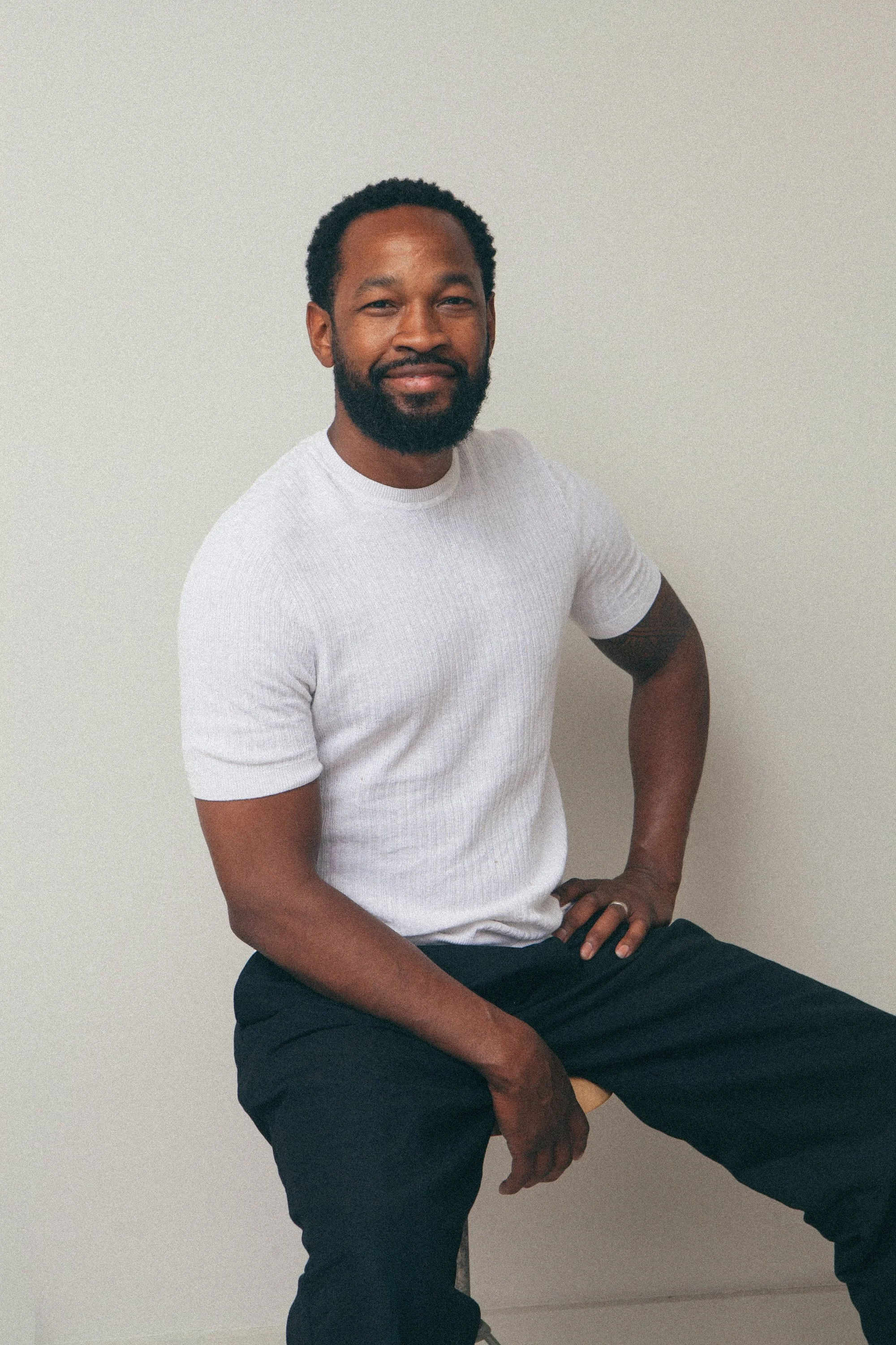 A smiling man with a beard and short hair, dressed in a dark green shirt, resting his arms on a surface in an outdoor setting.
