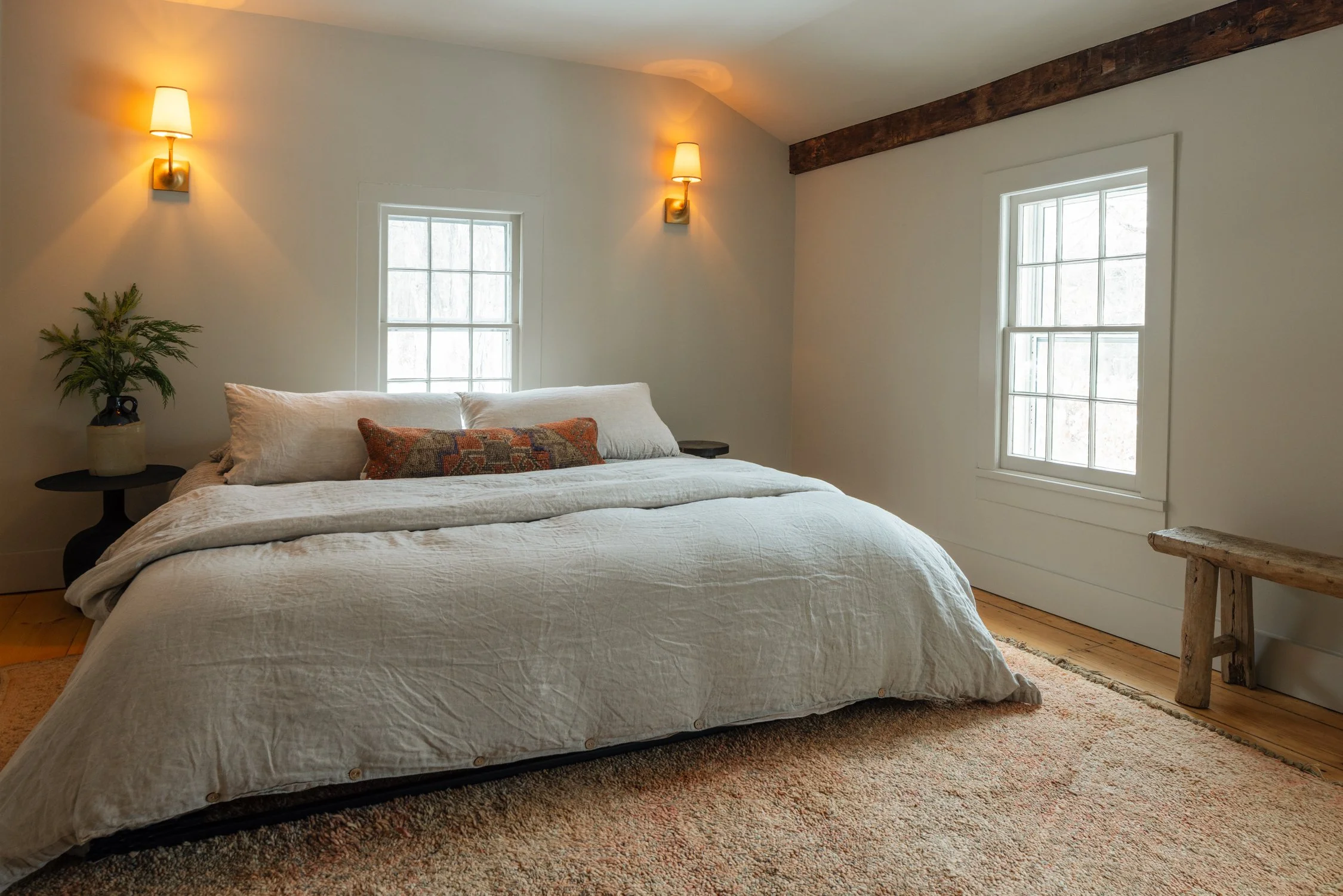 A cozy bedroom with a bed covered in a white quilt, two beige pillows, and a decorative pillow. Two windows bring in natural light. There are wall-mounted lamps on either side of the bed, a small side table with a plant on the left, and a rustic wood