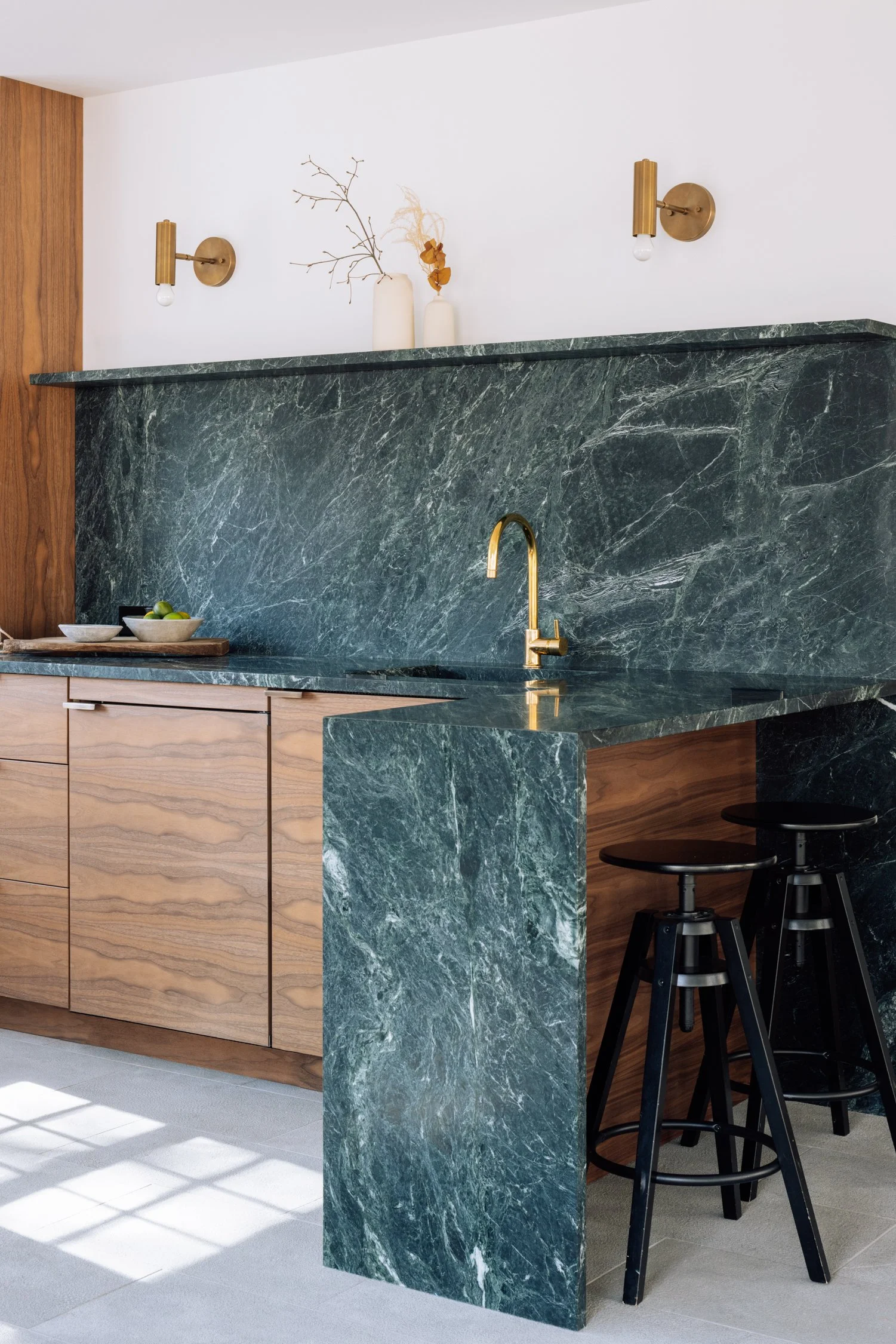 Modern kitchen with green marble countertops and backsplash, wooden cabinets, brass fixtures, and black bar stools.