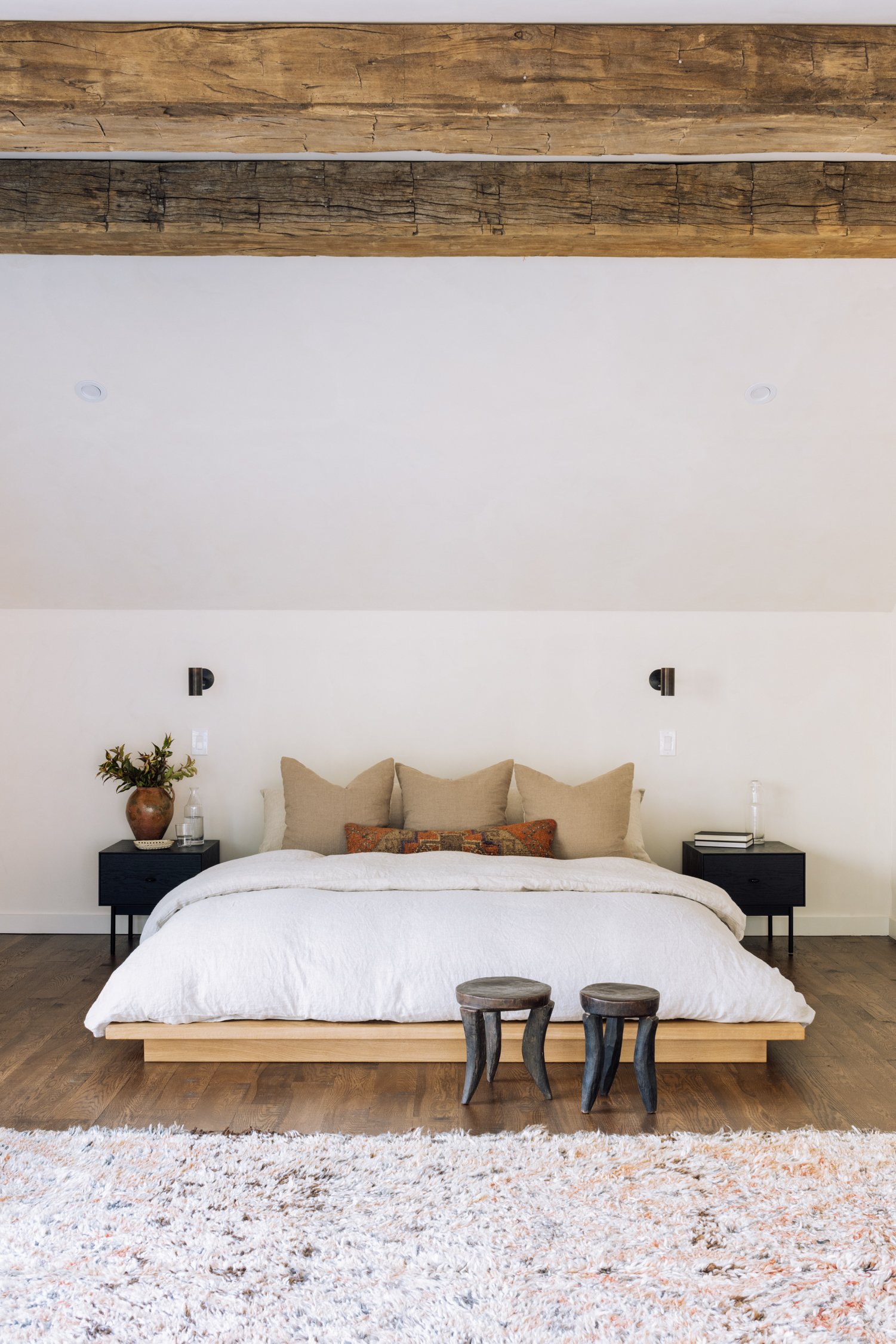 A modern bedroom with a large bed, beige pillows, black nightstands, wooden flooring, a textured white bedspread, a colorful pillow, two small wooden stools, a plush rug, and a wooden ceiling beam.