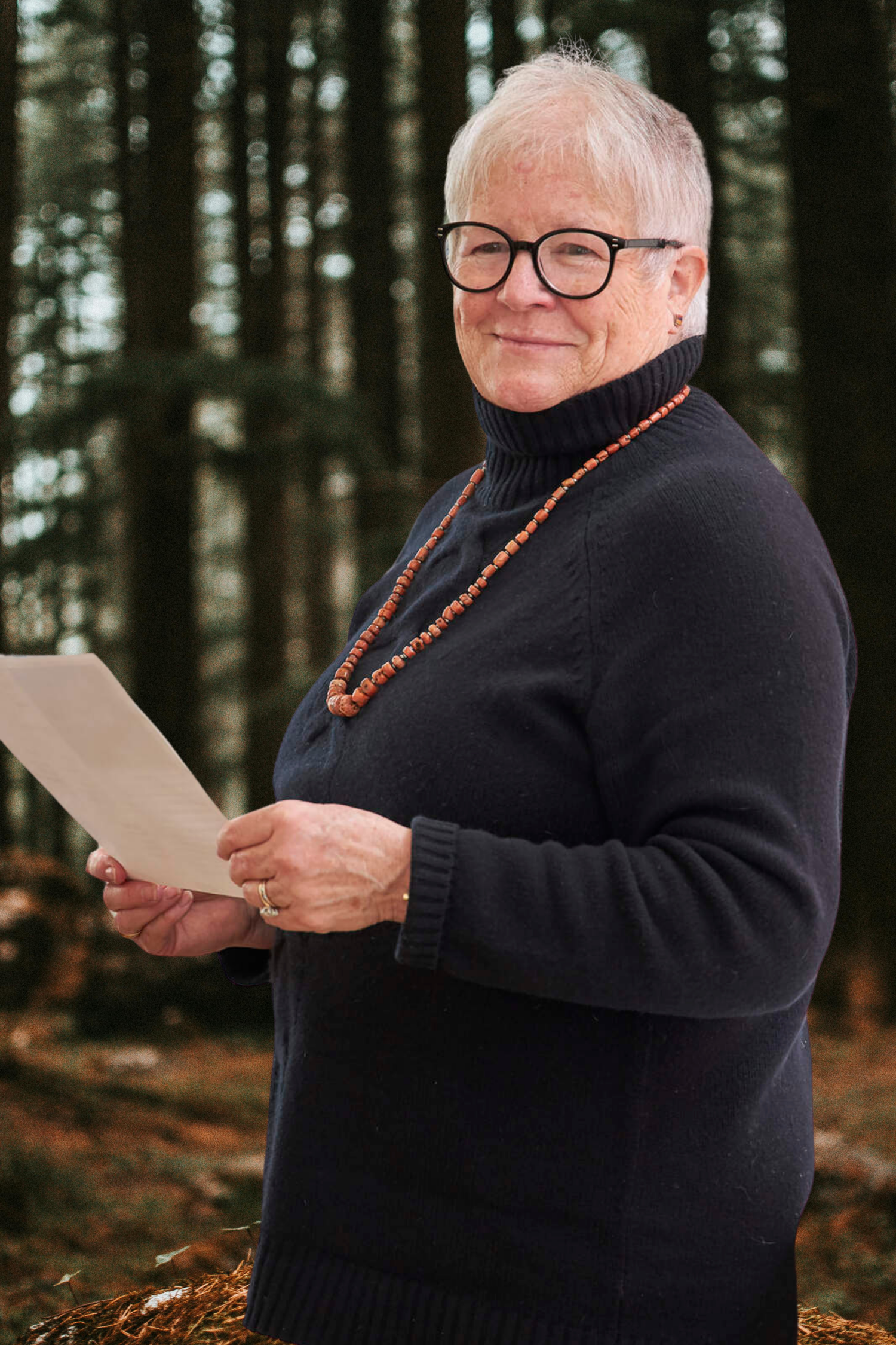 An elderly woman with short gray hair, glasses, and a black sweater with a turtleneck, holding a piece of paper and smiling, standing outdoors in a wooded area.