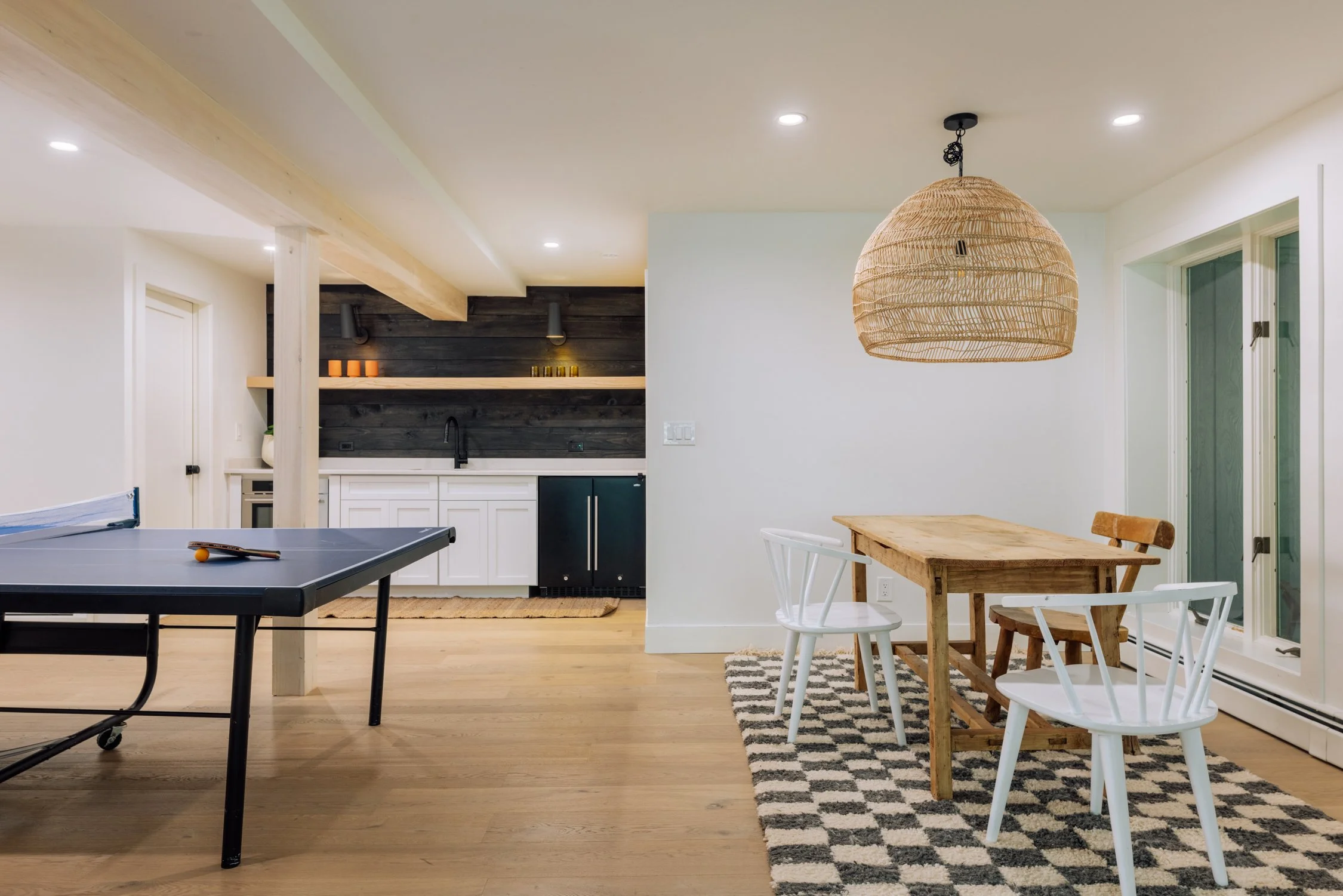 Indoor scene with a black ping pong table on the left, a kitchenette with white cabinets and a black countertop in the background, and a dining area on the right with a wooden table, white and wooden chairs, a patterned rug, and a large woven pendant