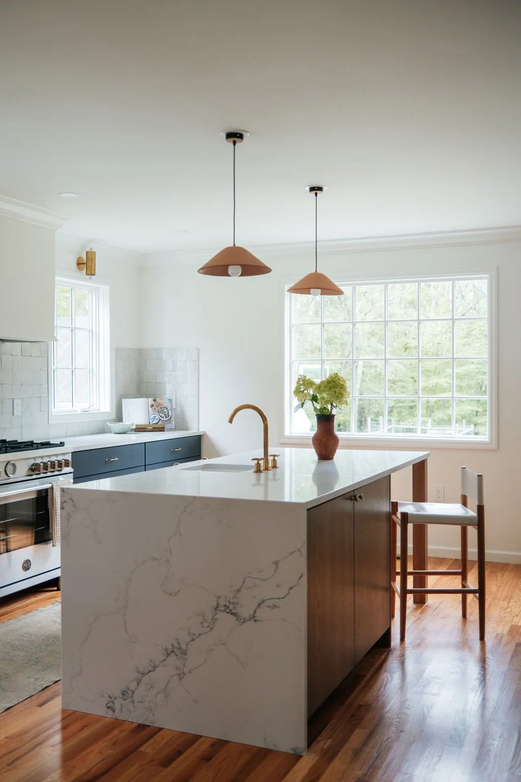 Modern kitchen with white marble island, wooden bar stool, blue cabinets, large window with greenery outside, pendant lights, and a vase with green and white flowers.
