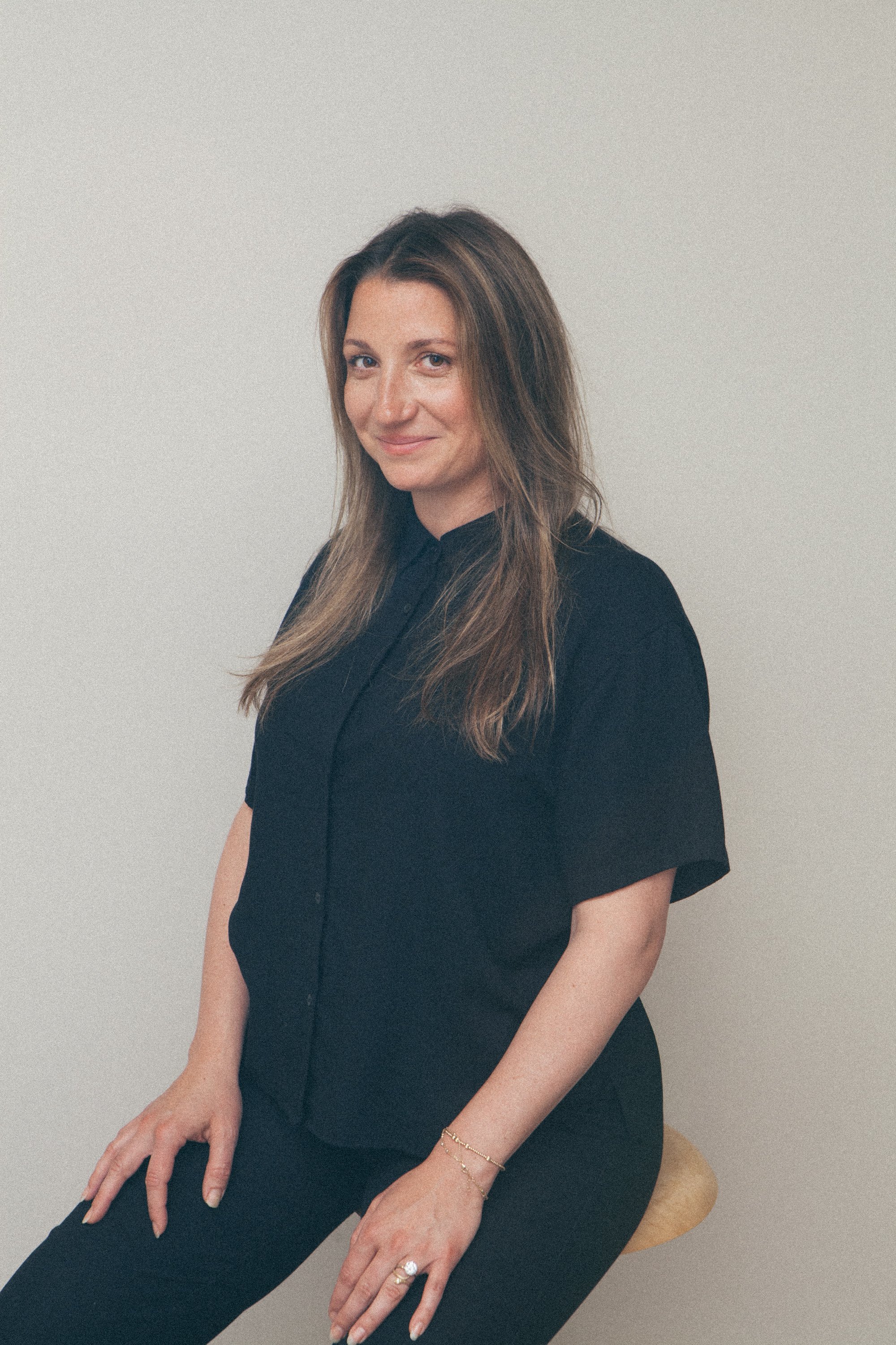 A woman with long brown hair and light skin, smiling softly at the camera, wearing a black jacket, indoors with a wooden background.