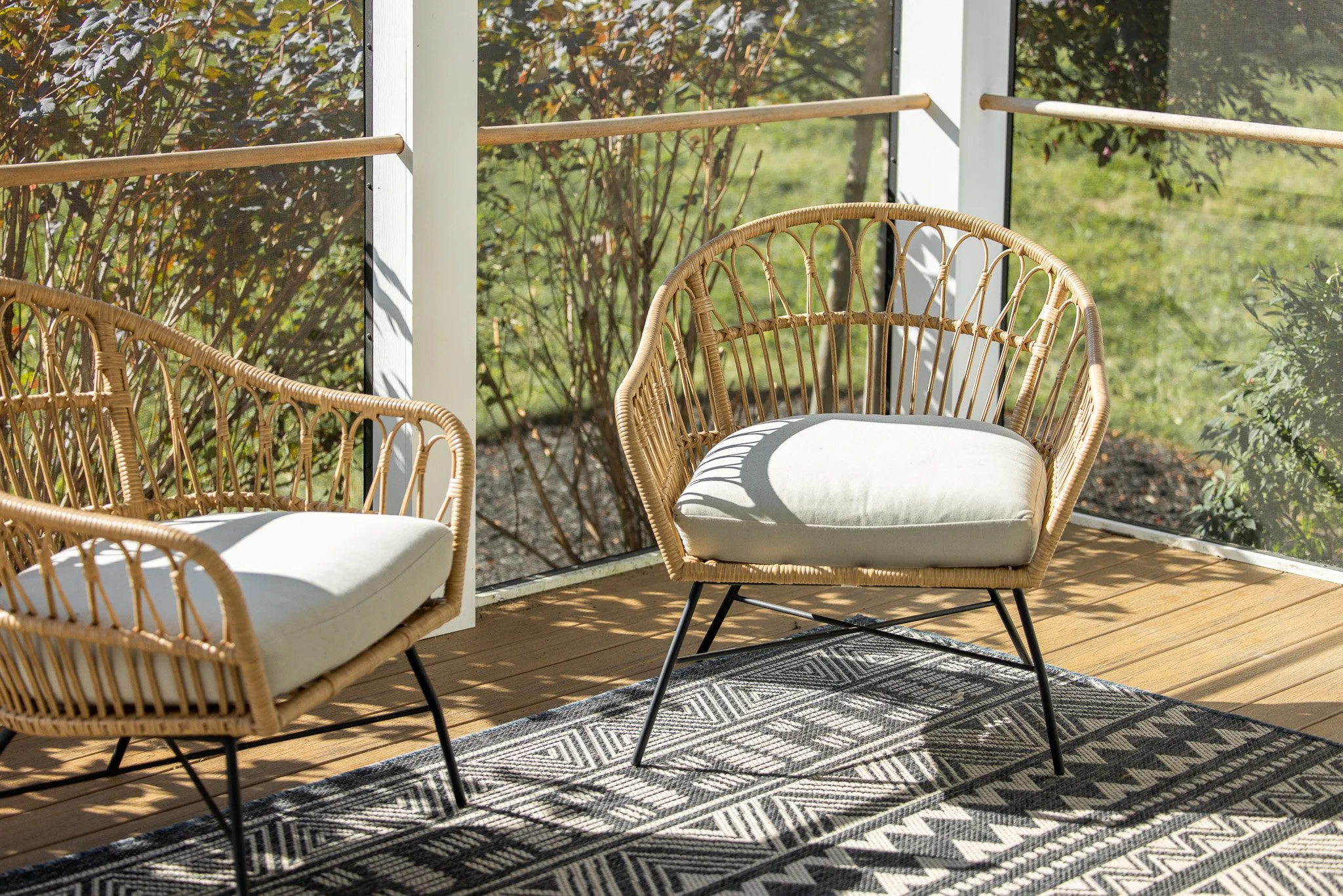 Sunlit porch with two rattan chairs with white cushions, black metal legs, and a patterned black and white rug, overlooking a garden with green grass and bushes.