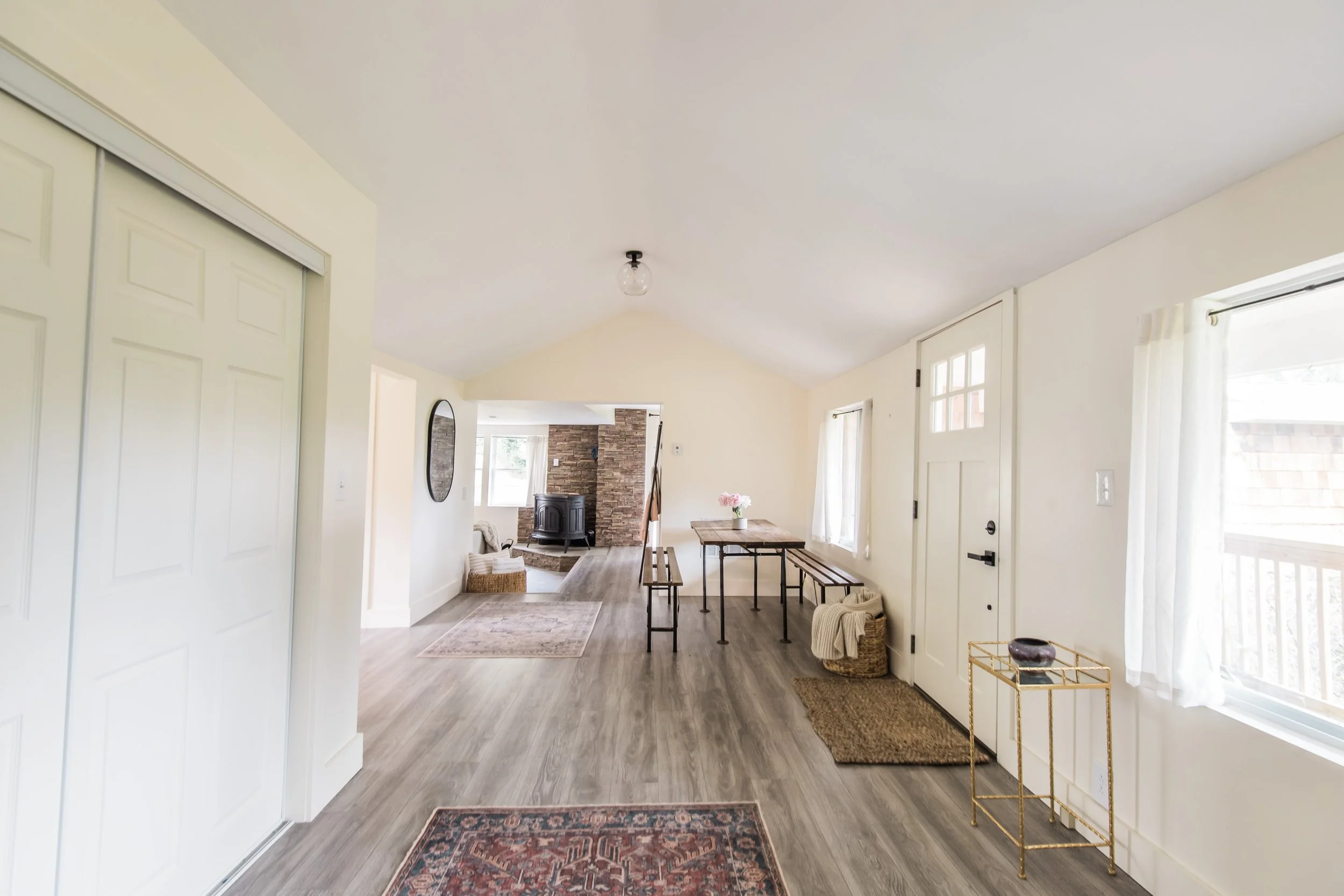 Living room with light-colored walls, wooden flooring, a table with benches, a wicker basket, a gold side table, and windows with white curtains.