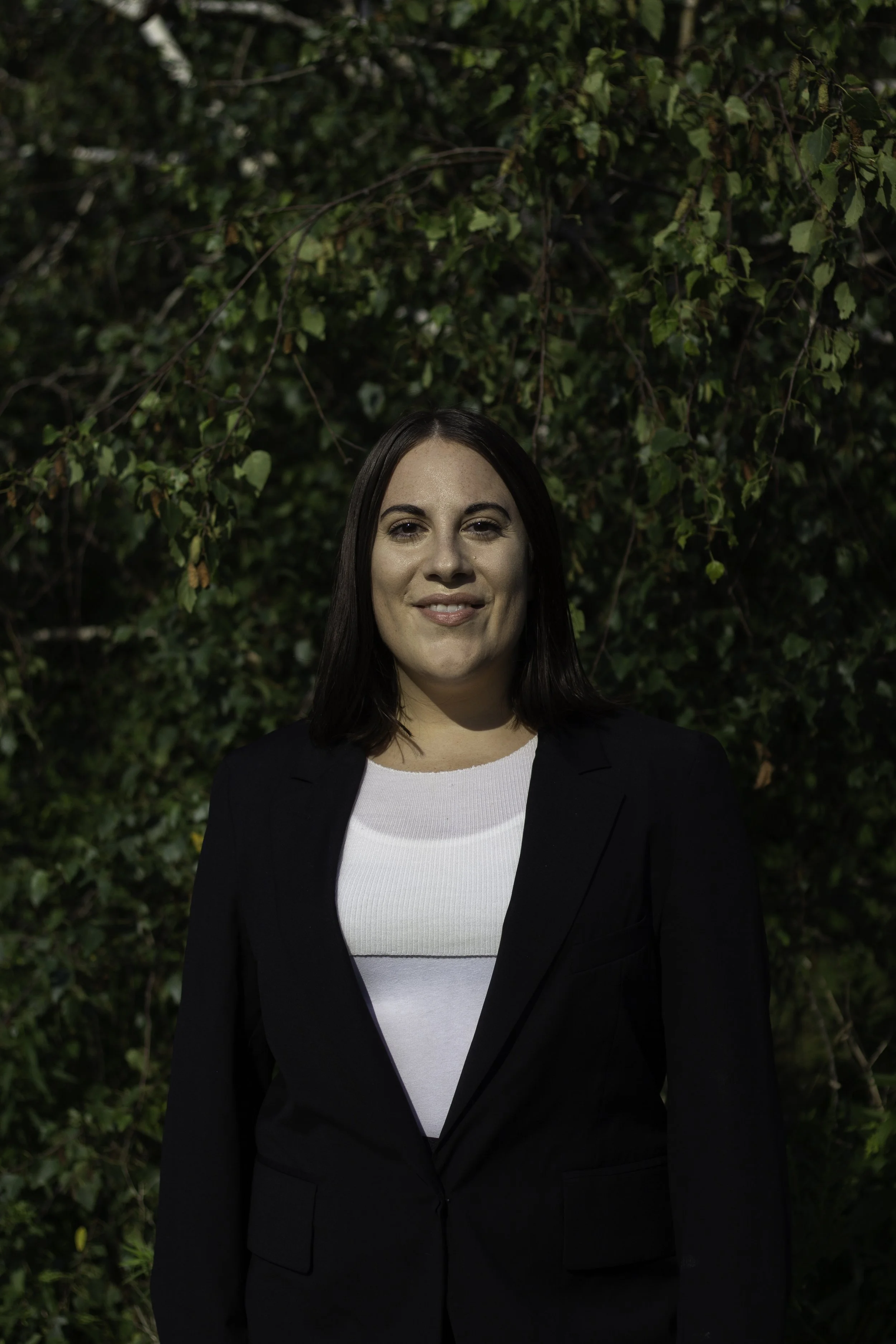 A woman with dark brown hair in a black blazer and white top standing outdoors in front of a dense green leafy background.