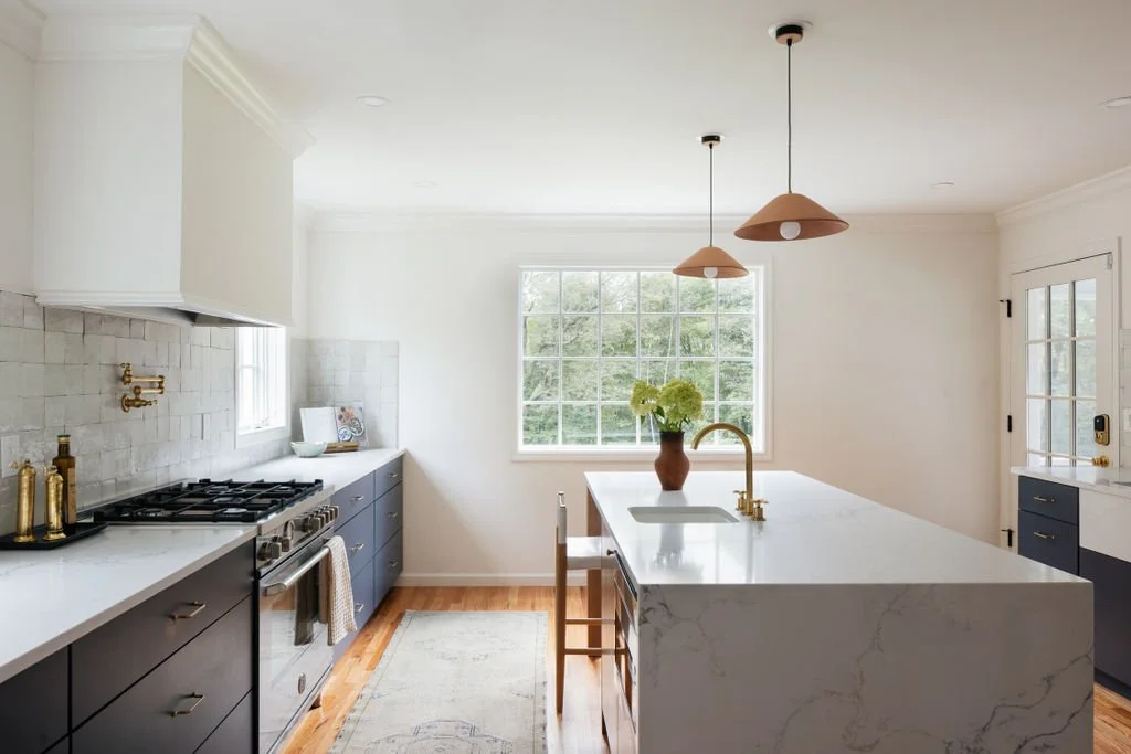 Modern kitchen with white walls, large window, marble island with gold faucet, navy lower cabinets, stainless steel stove, hardwood floor, pendant lights, and decorative bottles.