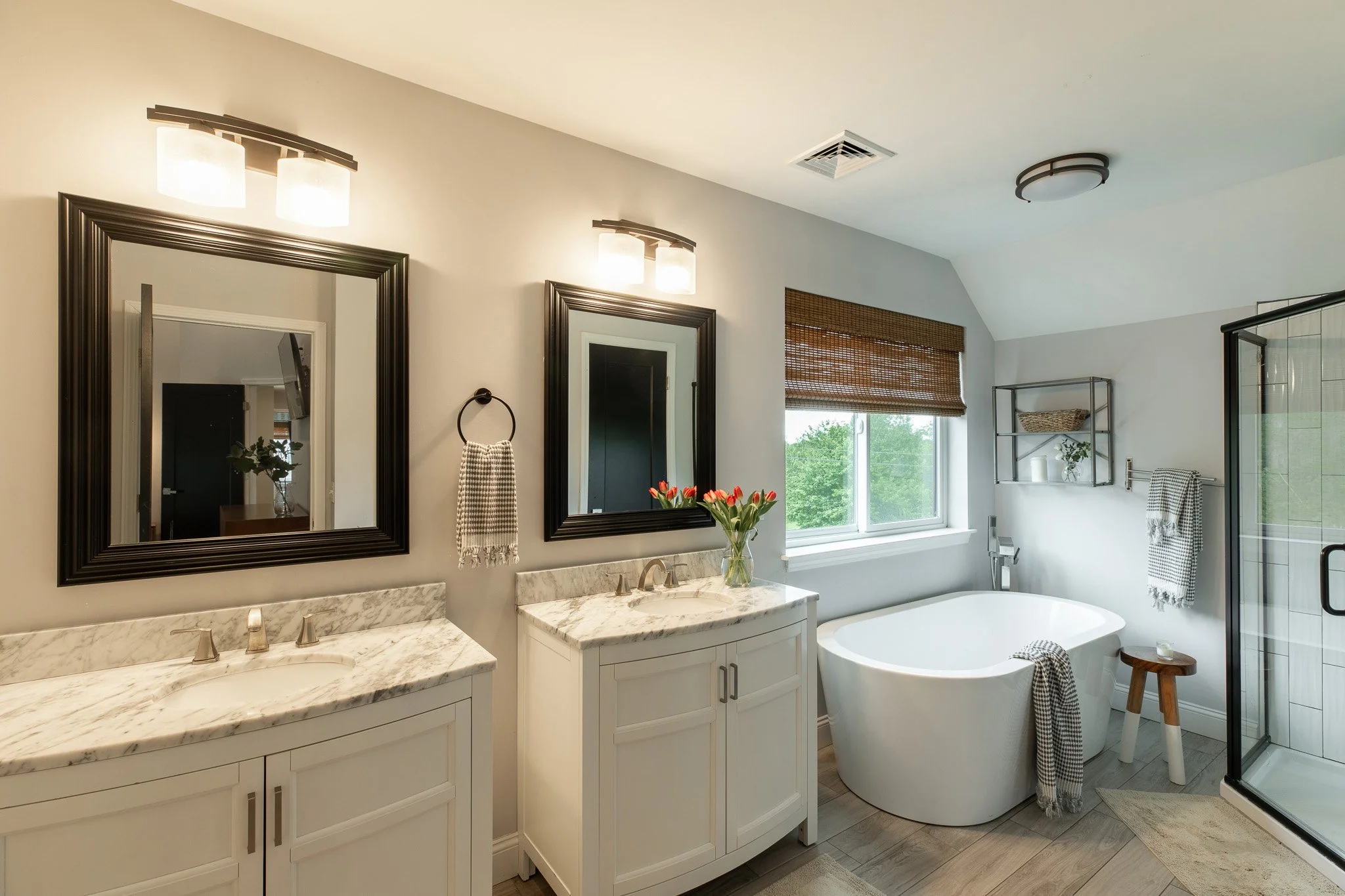 Modern bathroom with dual sinks, large mirrors, a freestanding bathtub, a glass-enclosed shower, and natural light from a window.