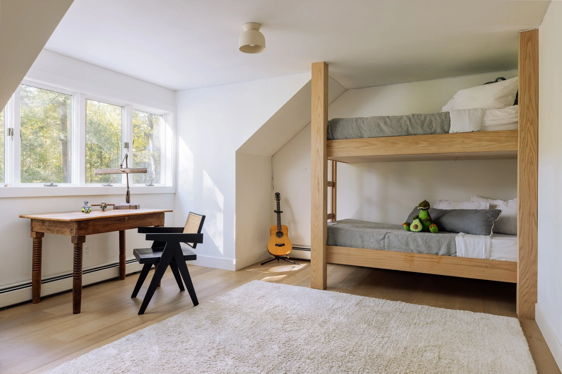 Room with a wooden bunk bed, a guitar on a stand, a desk near a window, and a plush toy on the lower bunk